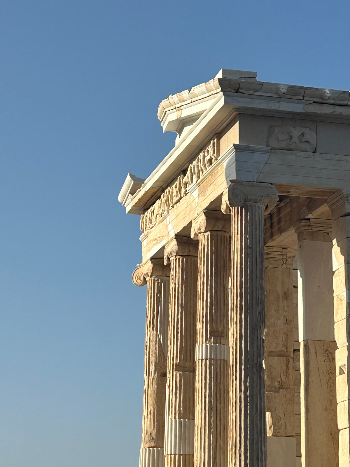Ancient Greek temple ruins with tall columns and sculpted friezes are shown in golden sunlight against a clear blue sky.