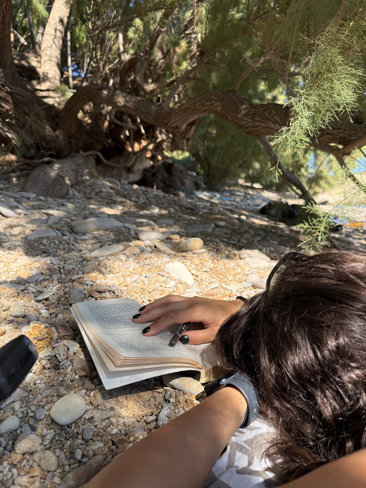 Person lying on rocky ground under tree shade, reading a book. Their hand with black-painted nails rests on the open pages. Sunlight filters through the branches overhead.