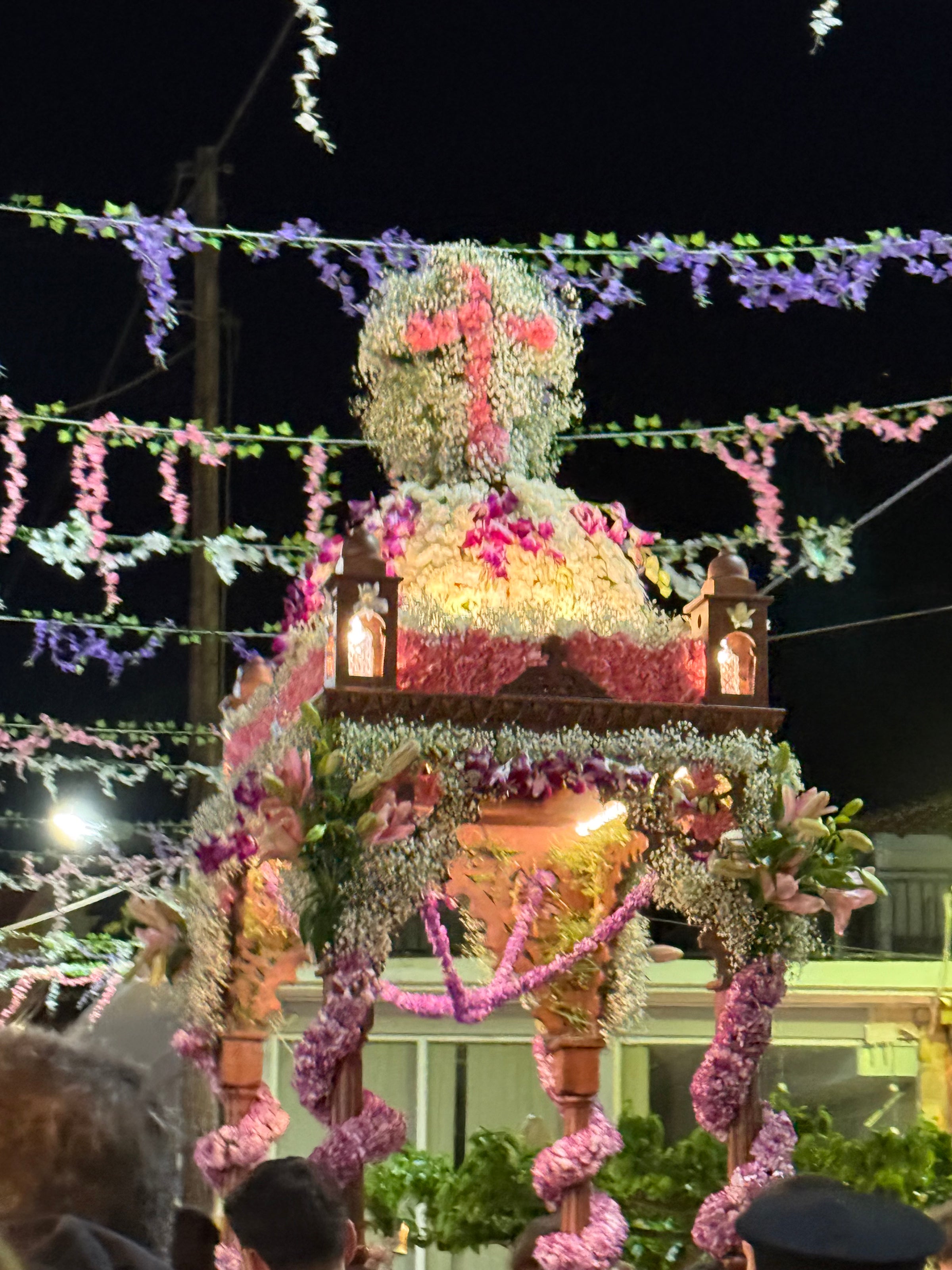 A large religious float covered in colorful flowers is carried through a nighttime street, surrounded by floral garlands and lights as people gather below.