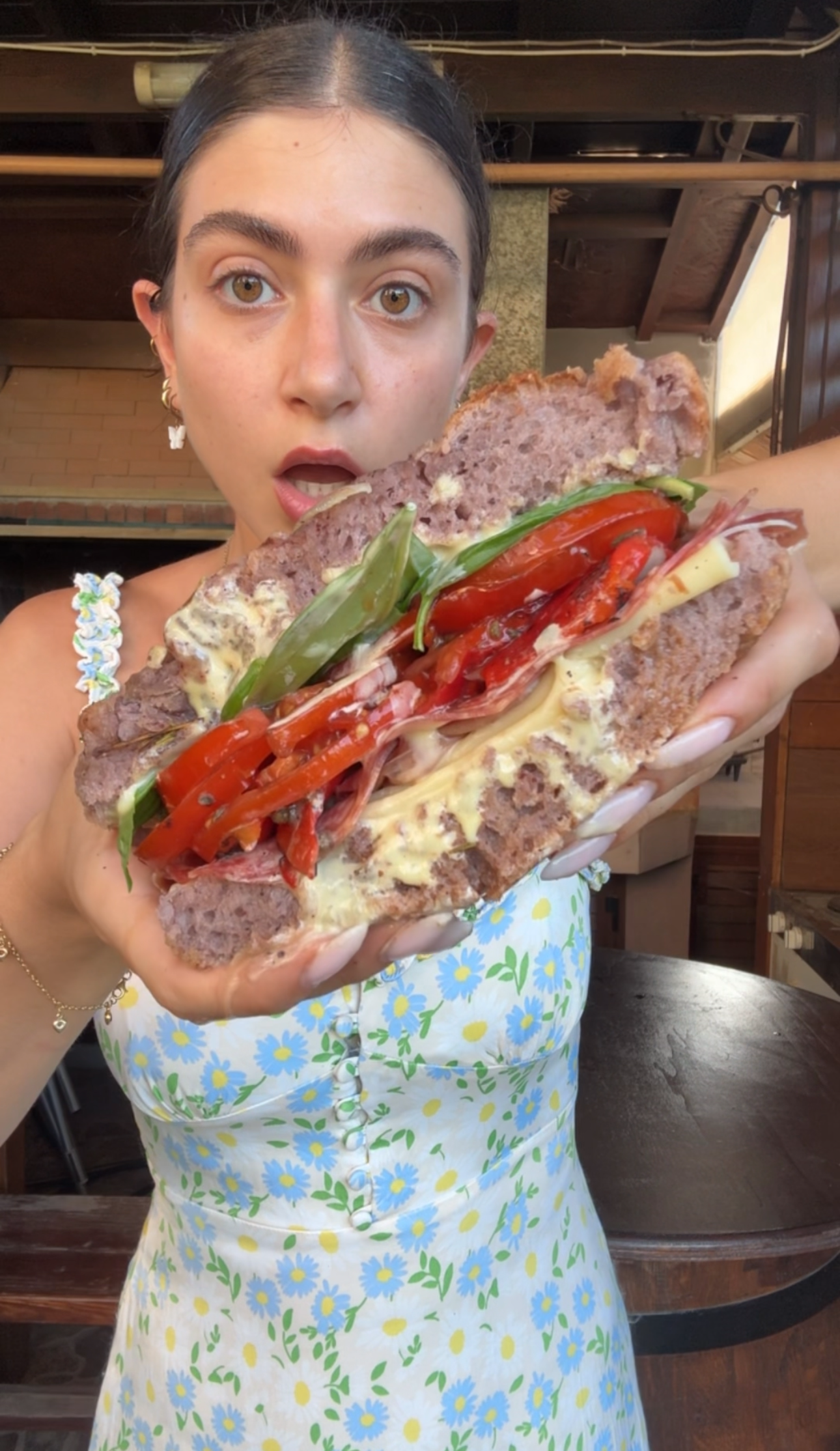 A woman in a floral dress holds a large sandwich filled with cheese, peppers, basil, and purple bread, looking surprised.