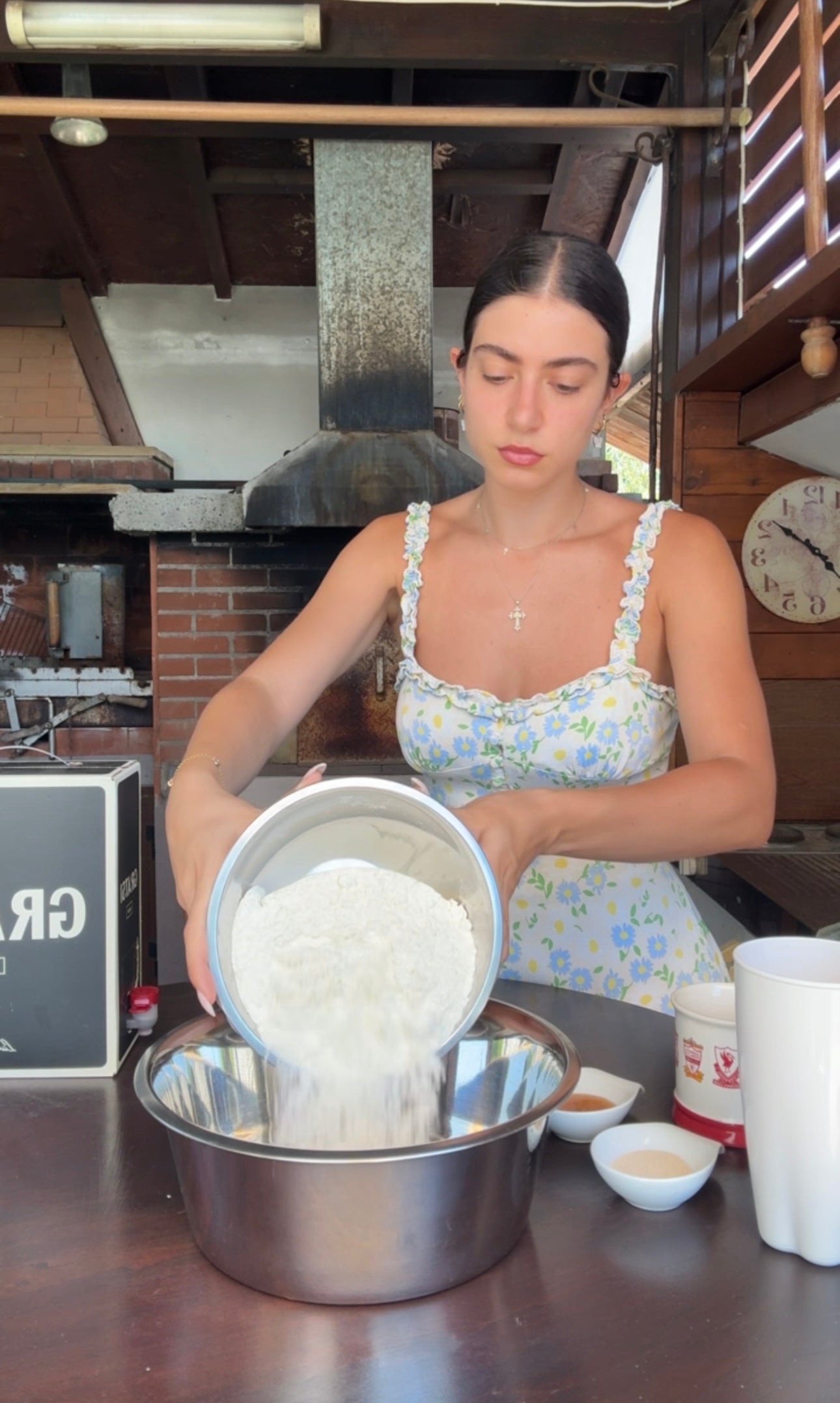 A woman in a floral dress pours flour from a container into a mixing bowl in a rustic kitchen with brick walls and wooden shelves.