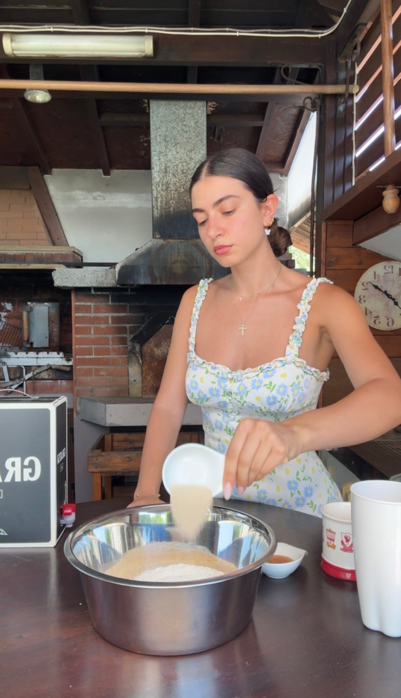 A woman in a floral dress pours sugar into a metal bowl in a rustic kitchen with a brick oven in the background.