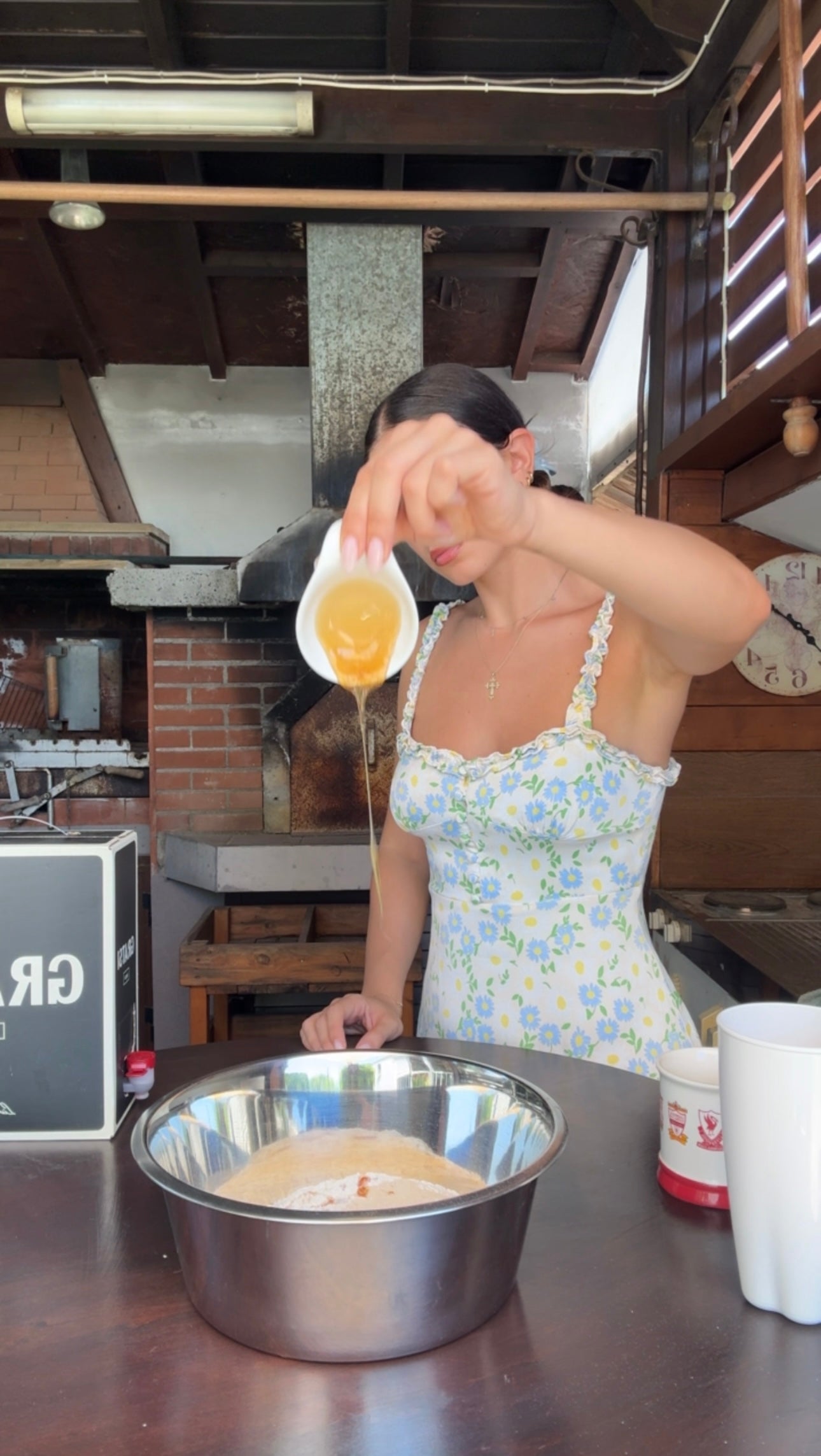 A woman in a floral dress pours honey from a small cup into a mixing bowl in a rustic kitchen setting.