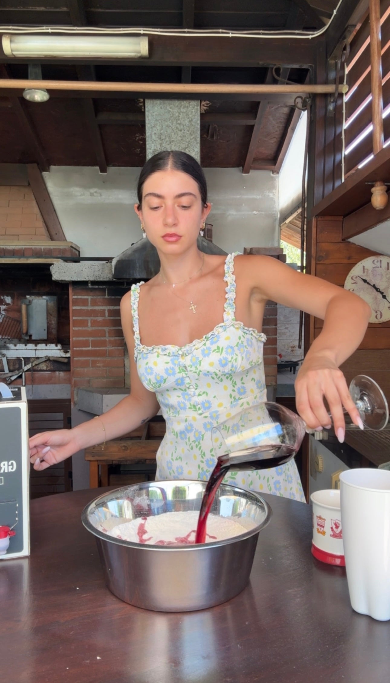 A woman in a floral dress pours red liquid from a glass into a metal mixing bowl in a rustic kitchen setting with brick and wood accents.