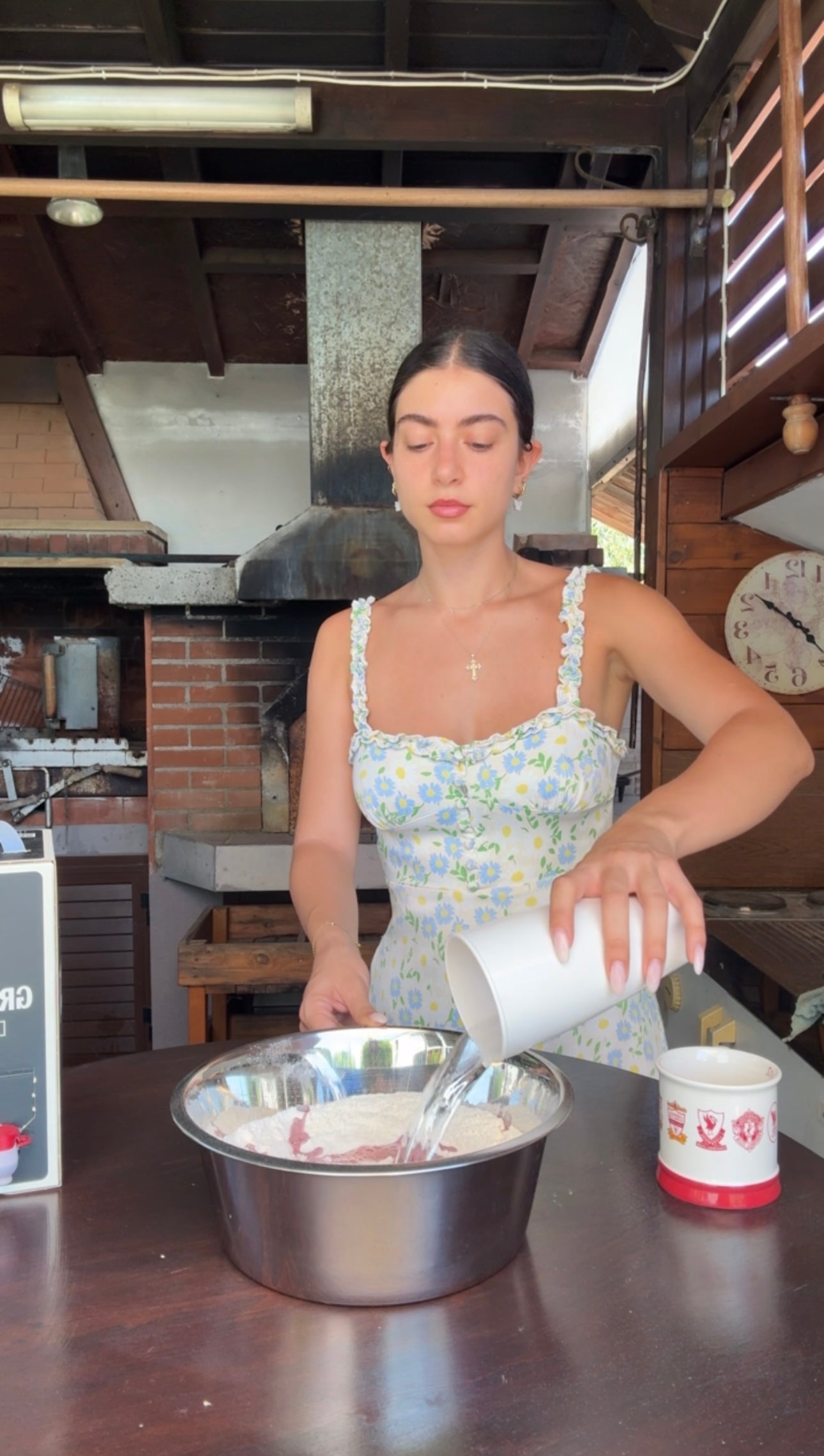A woman in a floral dress pours water into a metal bowl with flour, preparing to bake in a rustic kitchen with a brick oven in the background.