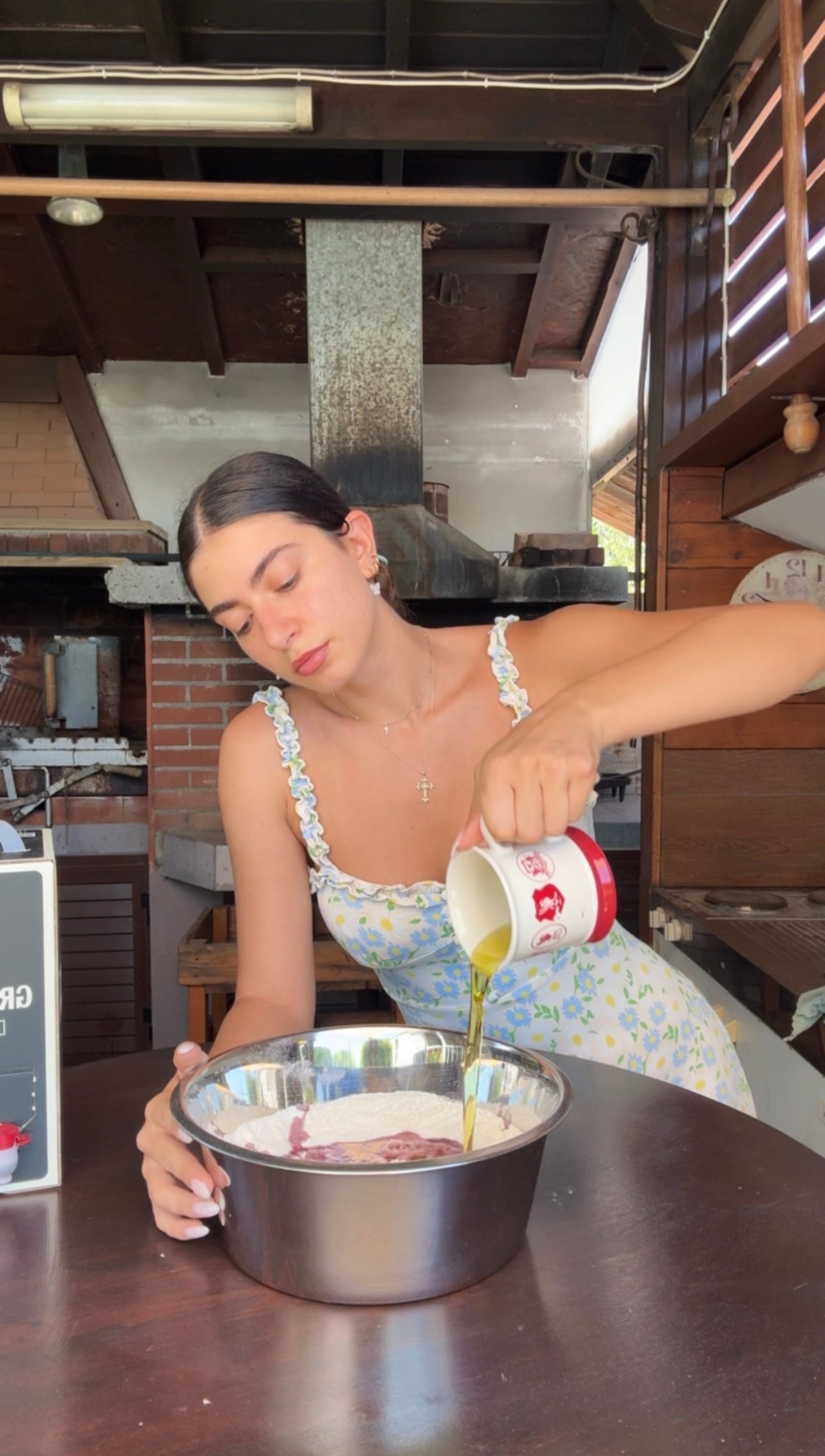 A woman in a floral dress pours olive oil from a cup into a bowl with flour at a wooden kitchen table in a rustic kitchen setting.