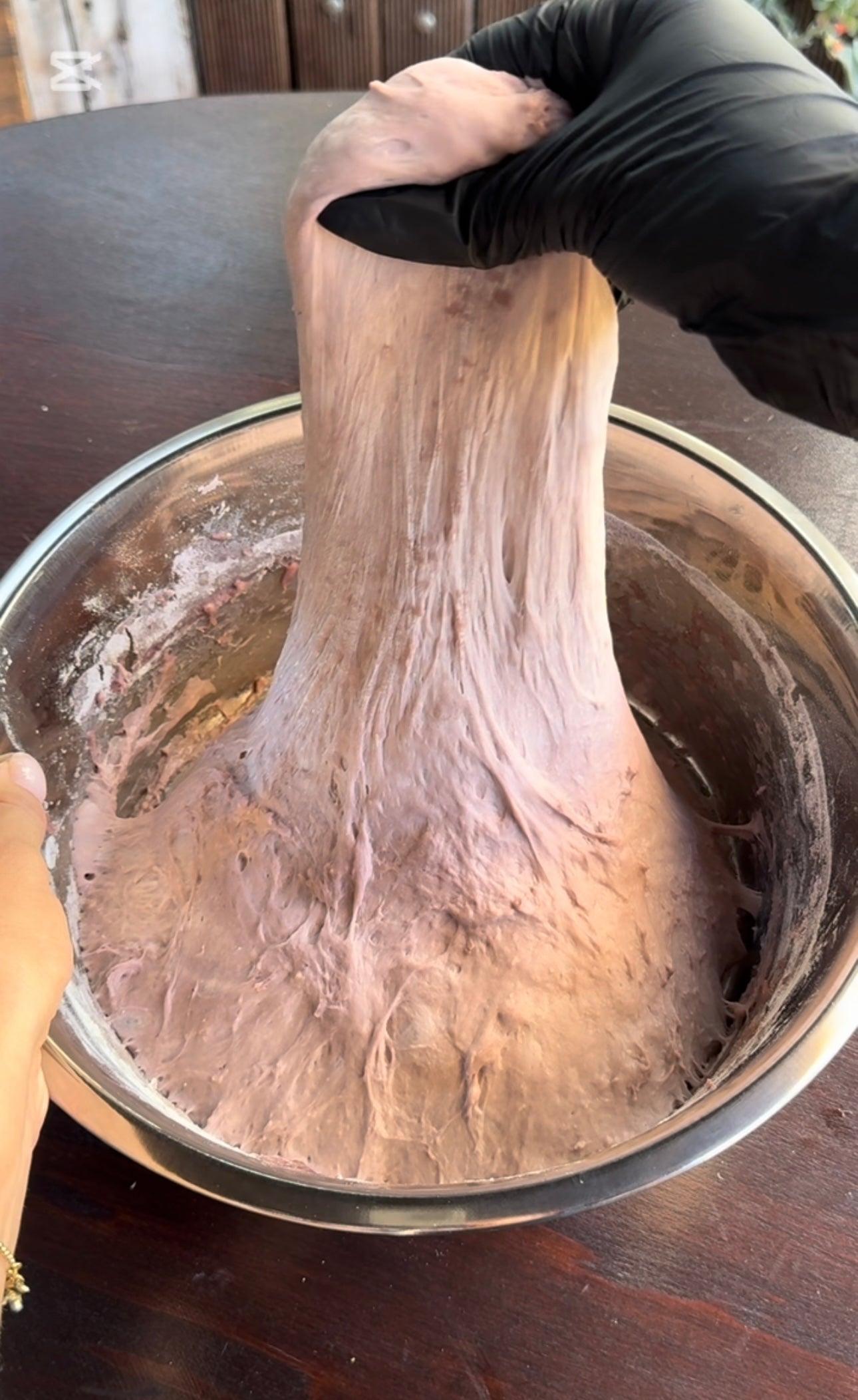 A stretchy, light purple dough is being lifted from a metal bowl by gloved hands, showing its sticky, elastic texture. Another hand holds the bowl steady on a dark wooden surface.
