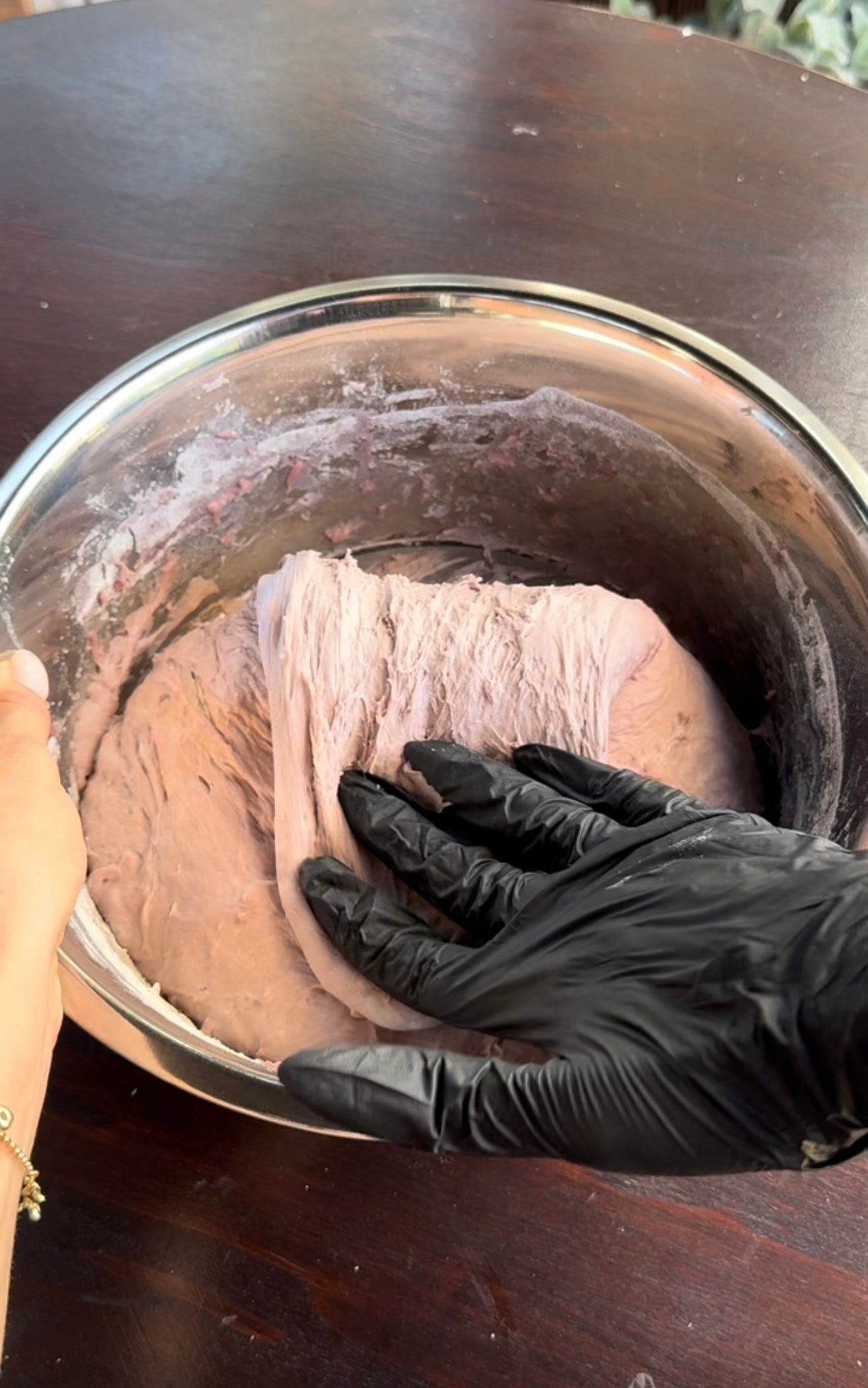 A hand in a black glove kneads light purple dough in a metal bowl, while another ungloved hand holds the bowl on a dark wooden surface.