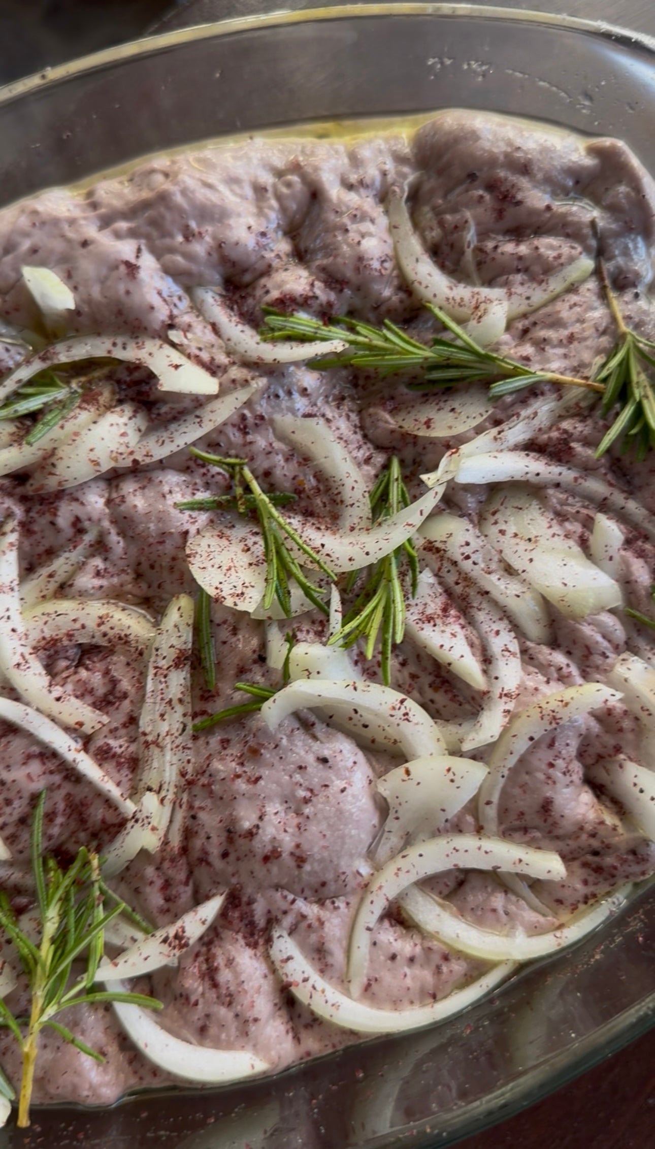 Raw marinated chicken pieces topped with sliced onions, fresh rosemary sprigs, and sprinkled with sumac, ready for cooking in a glass dish.