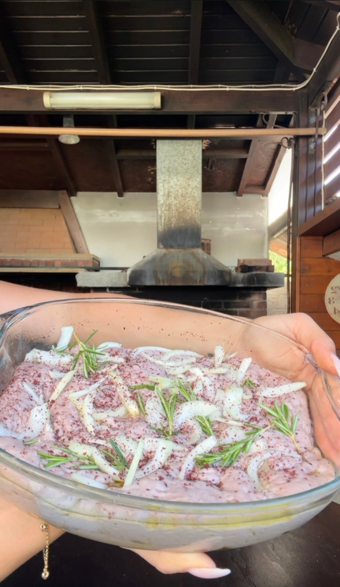 A person holds a glass dish filled with raw, seasoned chicken, onions, and rosemary, in an outdoor kitchen with a pizza oven in the background.