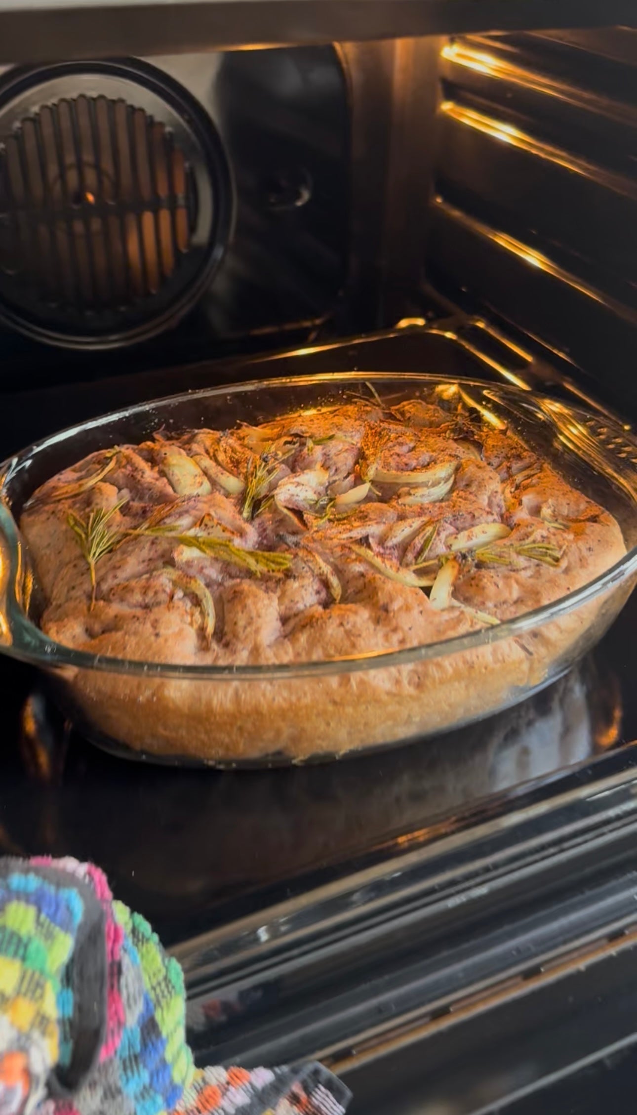 A glass baking dish with freshly baked focaccia bread topped with rosemary and onion sits on an oven rack, with a colorful towel visible at the edge.