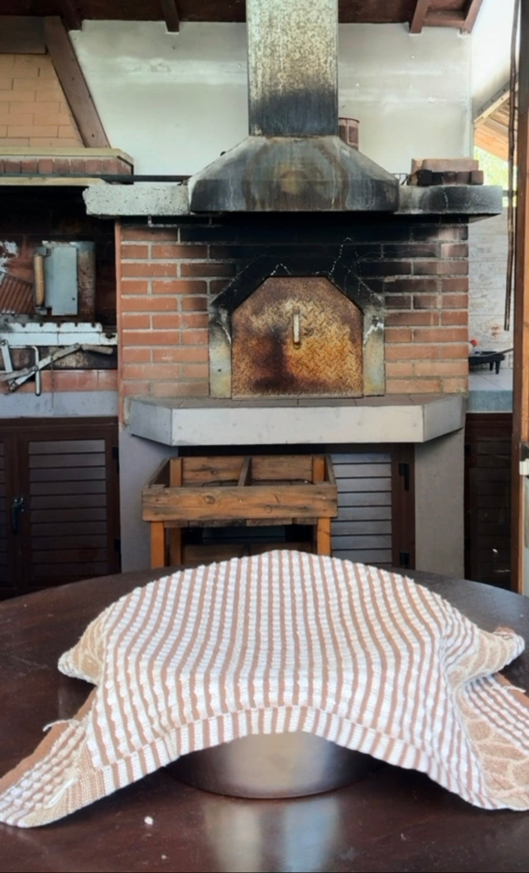 A metal bowl covered with a striped kitchen towel sits on a table in front of a rustic brick oven and chimney in a cozy kitchen setting.