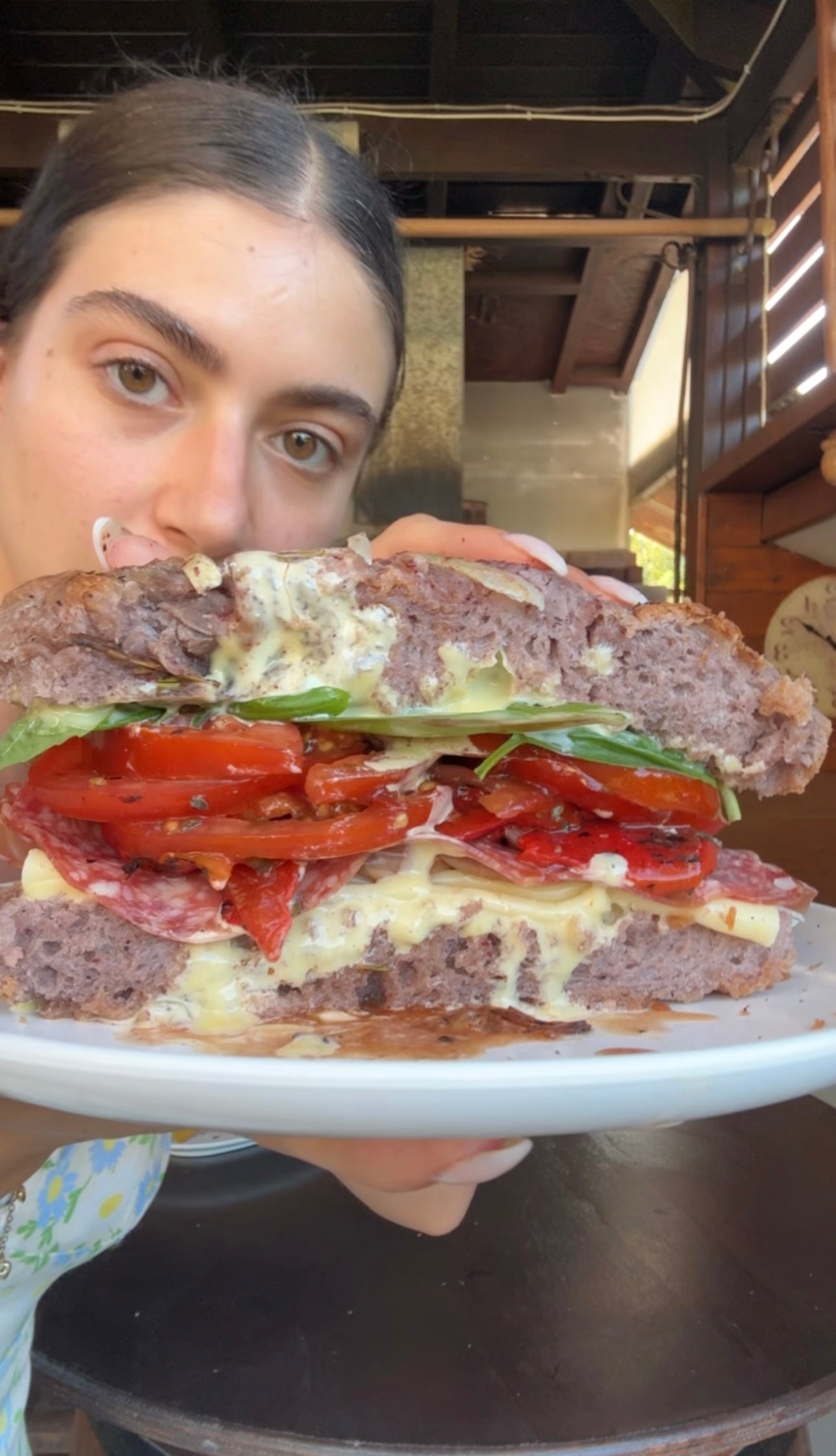 A person holds a large sandwich filled with melted cheese, roasted red peppers, greens, and salami on purple bread, close to their face.