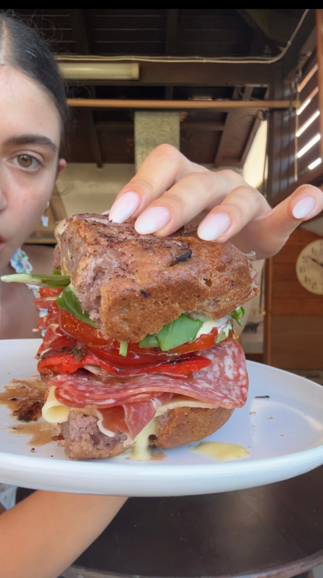 A person holds a large sandwich with salami, cheese, arugula, and roasted red peppers on rustic bread, on a white plate.