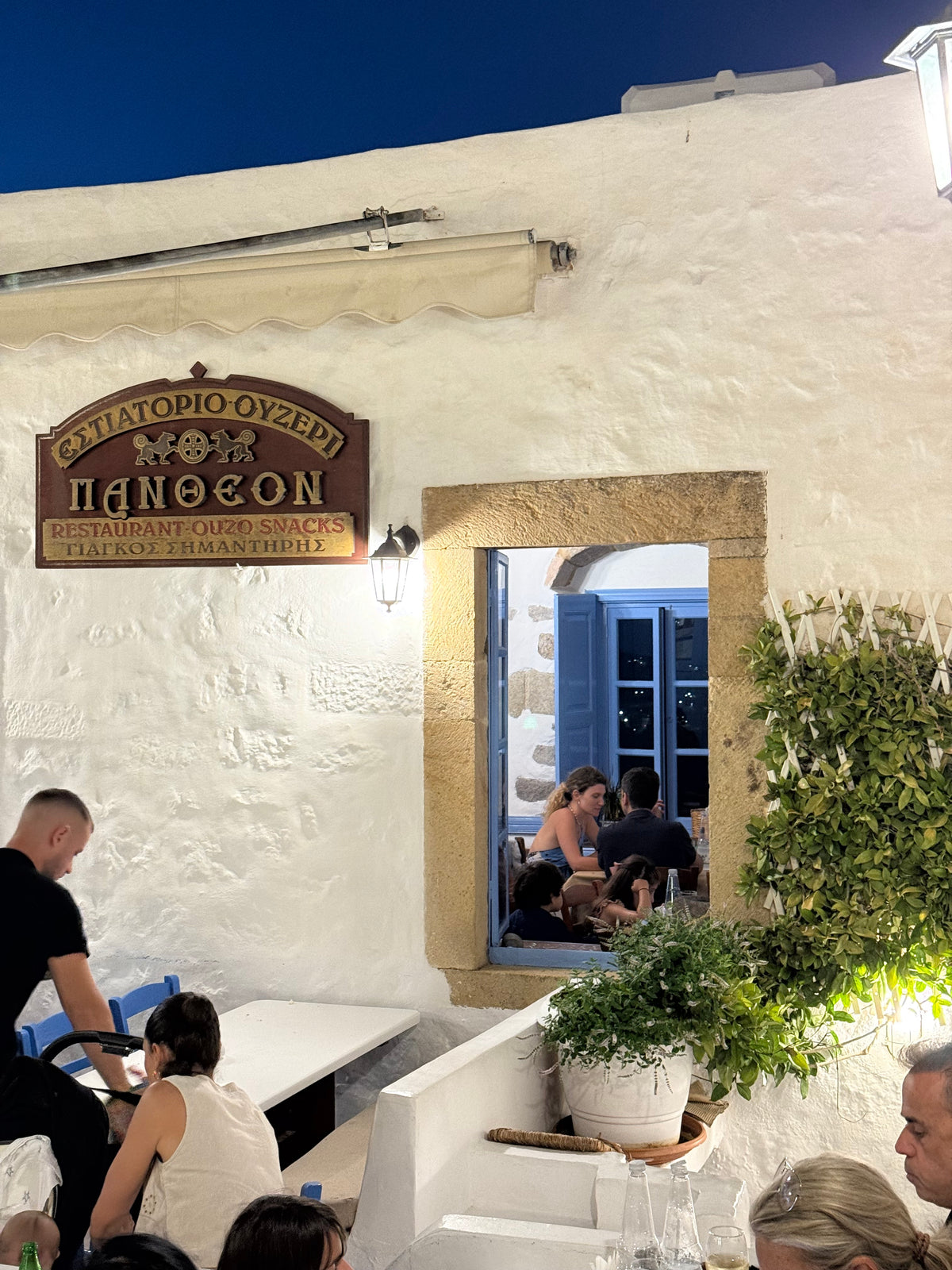 A bright restaurant with white walls and a window showing people dining inside. A sign in Greek and English hangs above the window, and potted plants decorate the scene. A few people are visible at tables outside.