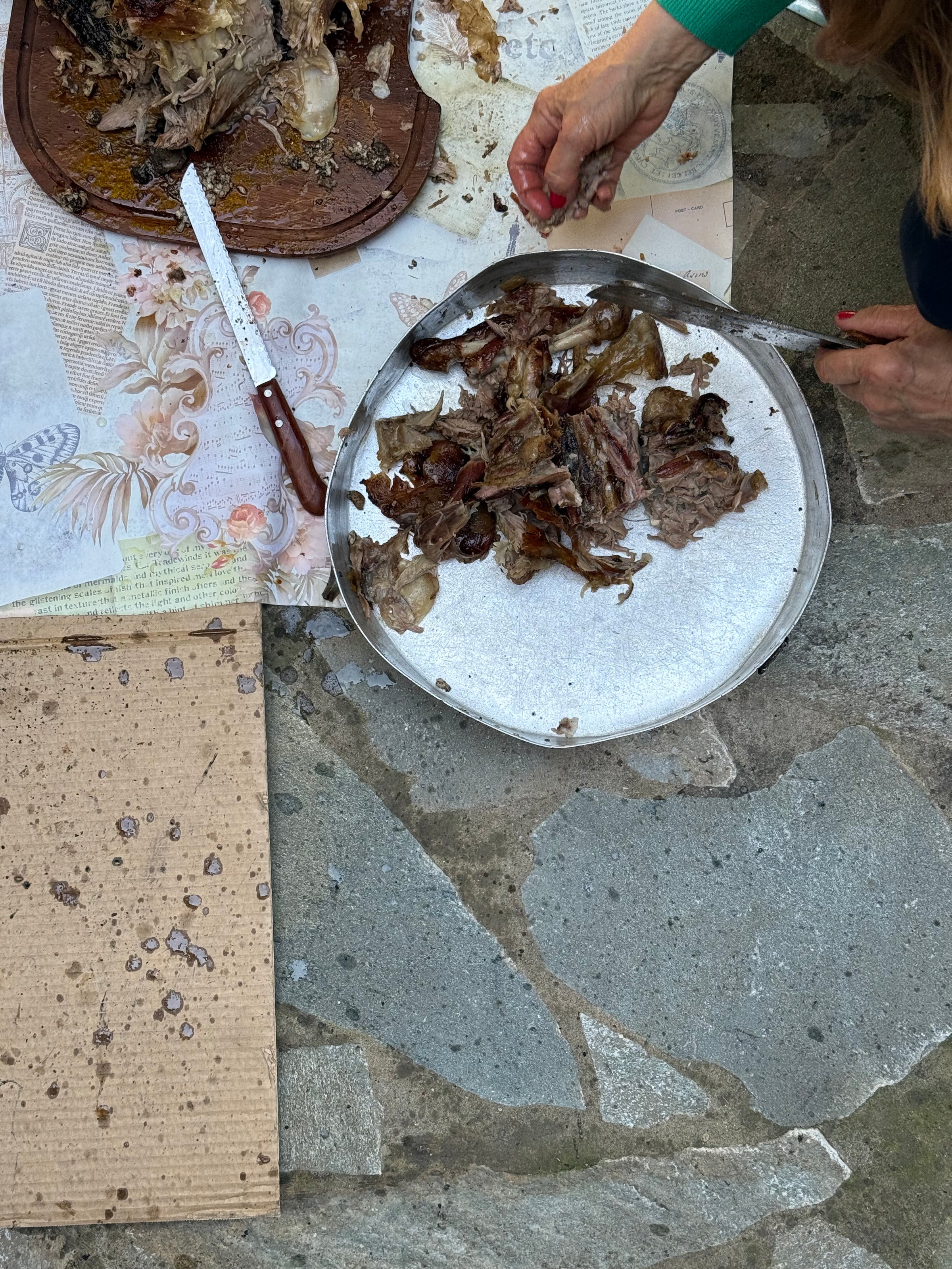 A person cuts cooked meat, placing pieces on a round tray on a stone floor. A knife rests on a wooden board with more meat and a floral-patterned paper nearby.