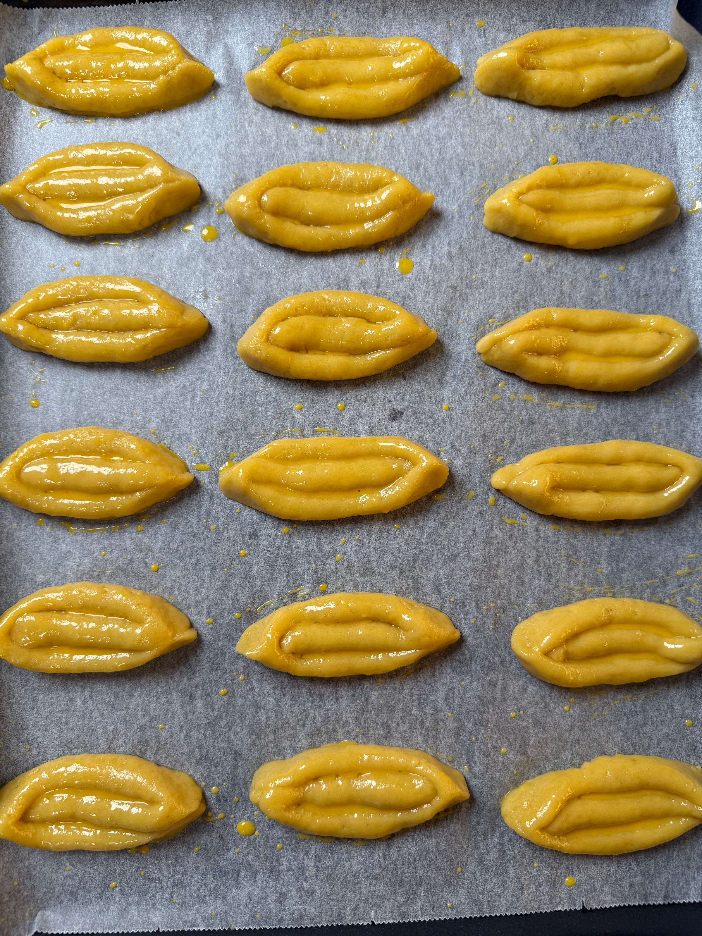Rows of yellow, oval-shaped pastries are arranged on a parchment-lined baking tray. The pastries have a glossy surface, likely from an egg wash, and feature a line indentation down the center.