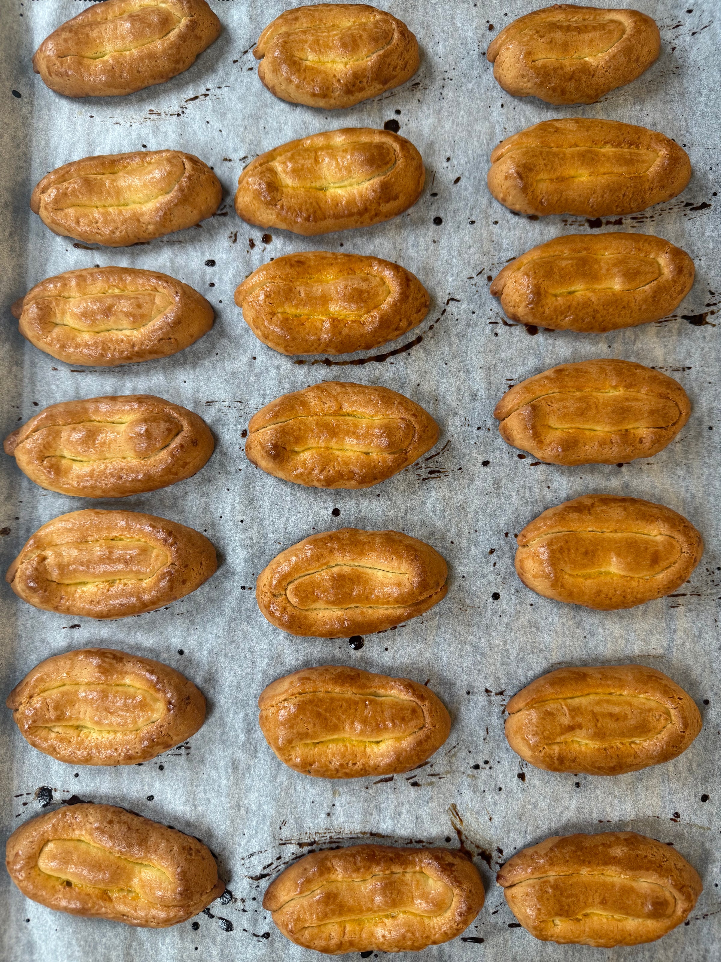 A baking tray lined with parchment paper holds twenty golden-brown, oval-shaped pastries arranged in neat rows. The pastries have a shiny, glazed surface and visible score marks on top.