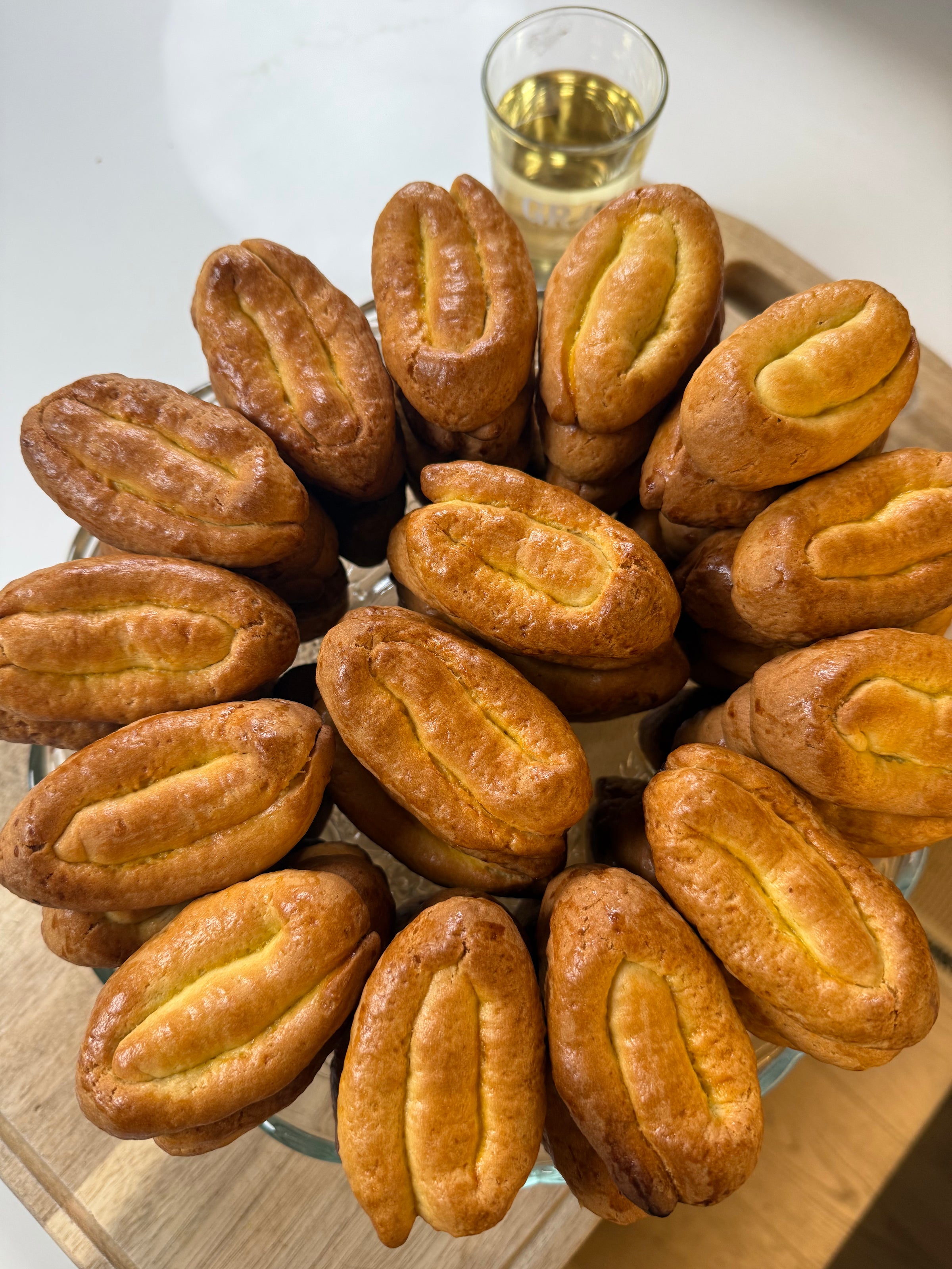 A glass plate filled with neatly arranged, oval-shaped, golden-brown pastries, each with a split top. In the background, there is a small glass of light yellow liquid on a wooden surface.