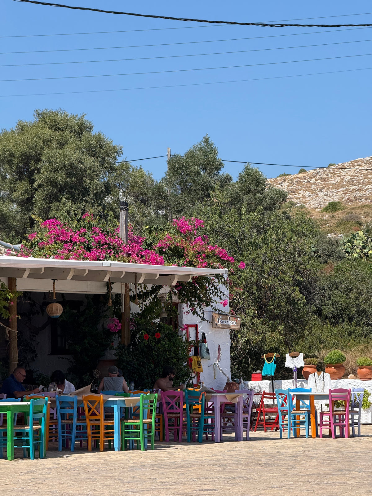 Colorful chairs and tables sit outside a quaint café with pink flowers overhead, surrounded by greenery and hills under a clear blue sky.