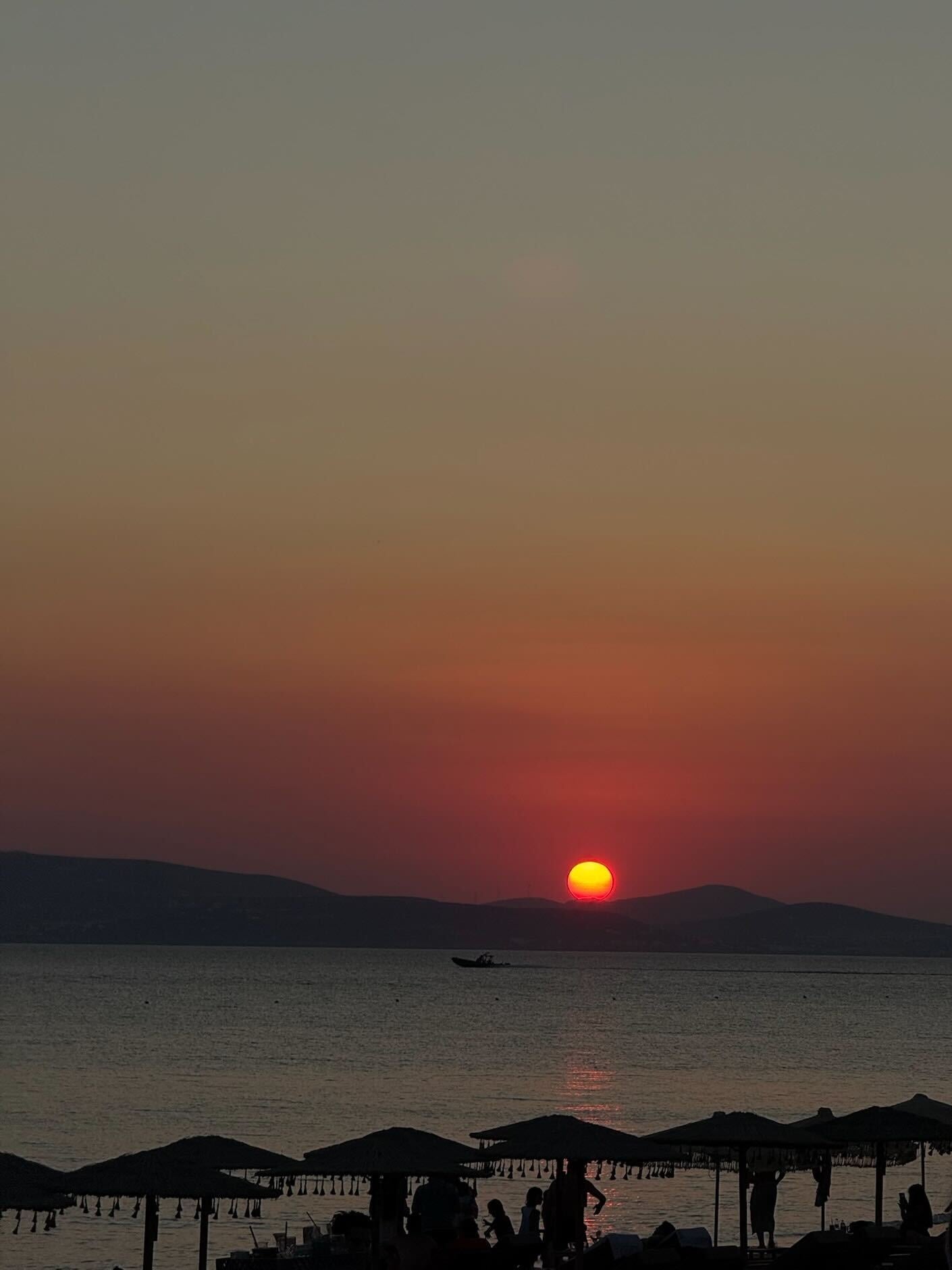 A vibrant sunset over the sea, with the sun low on the horizon. Silhouetted umbrellas and people are visible in the foreground, and a boat floats on the calm water against distant dark hills.