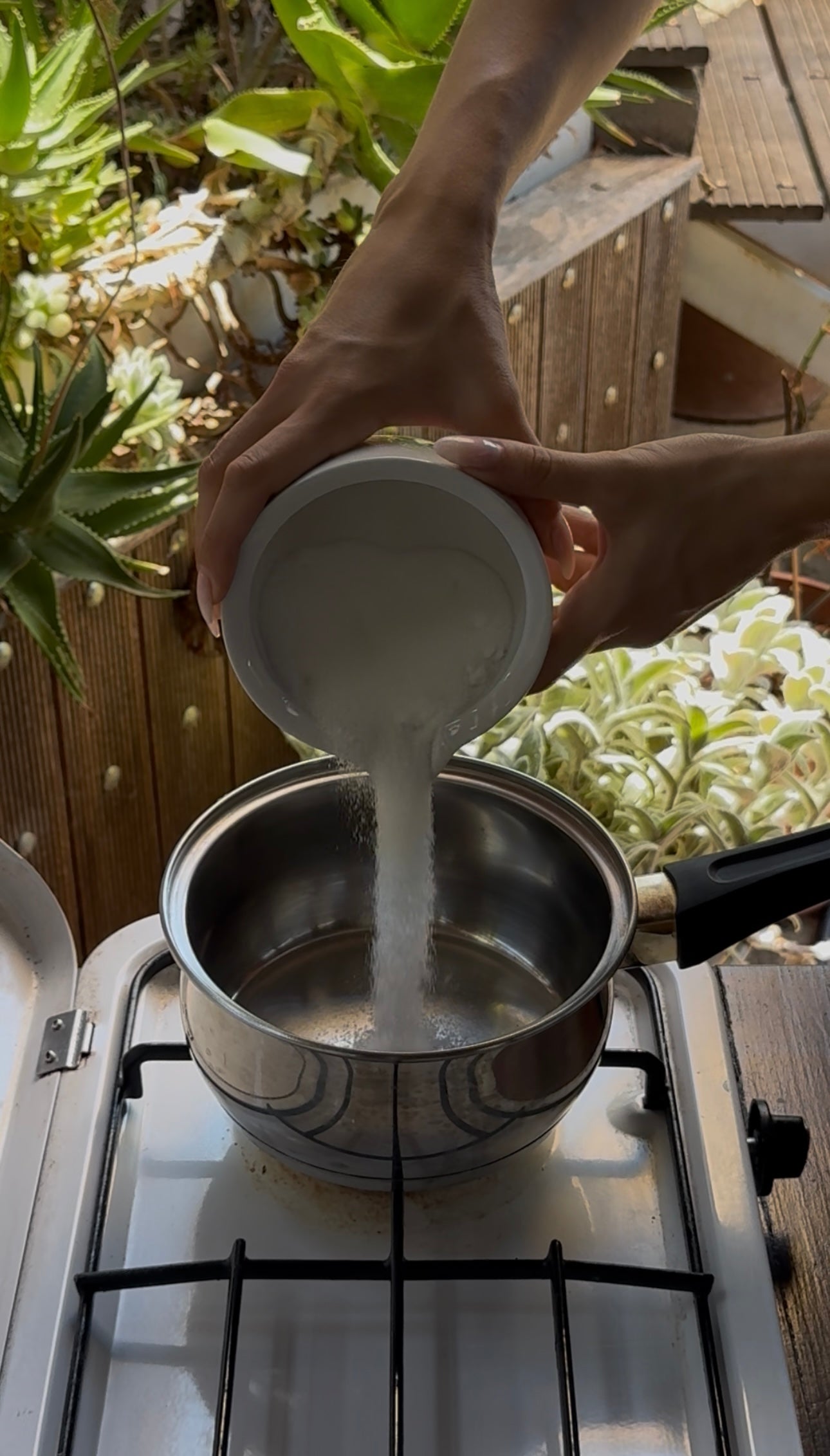 A person pours granulated sugar from a white bowl into a pot on a gas stove, with potted plants in the background.