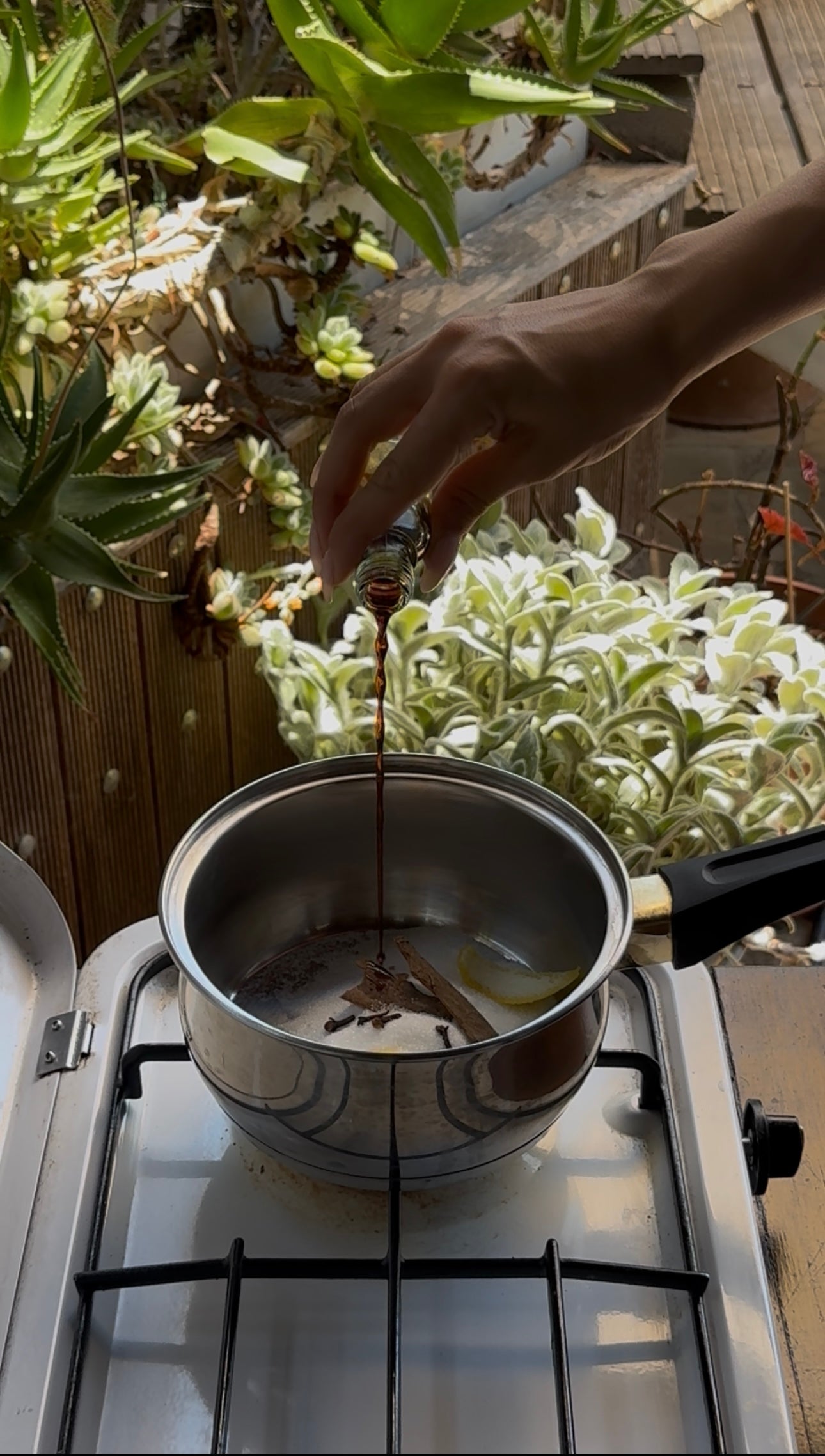A hand pours a dark liquid from a small bottle into a stainless steel pot on a gas stove, with green and white leafy plants in the background.