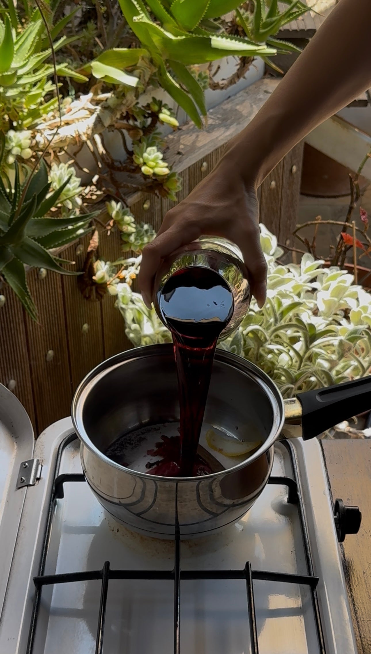 A hand pours a dark red liquid from a glass cup into a saucepan on a stove, with green plants in the background.
