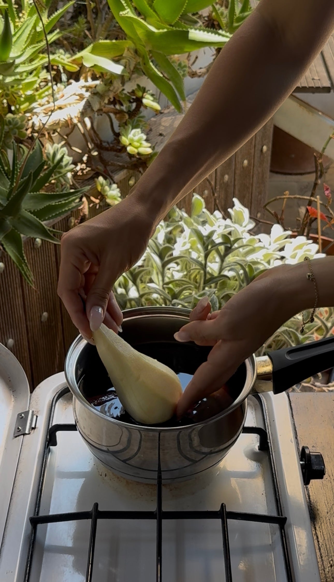 A person dips a peeled pear into a pot of dark liquid on a stove, surrounded by potted plants.