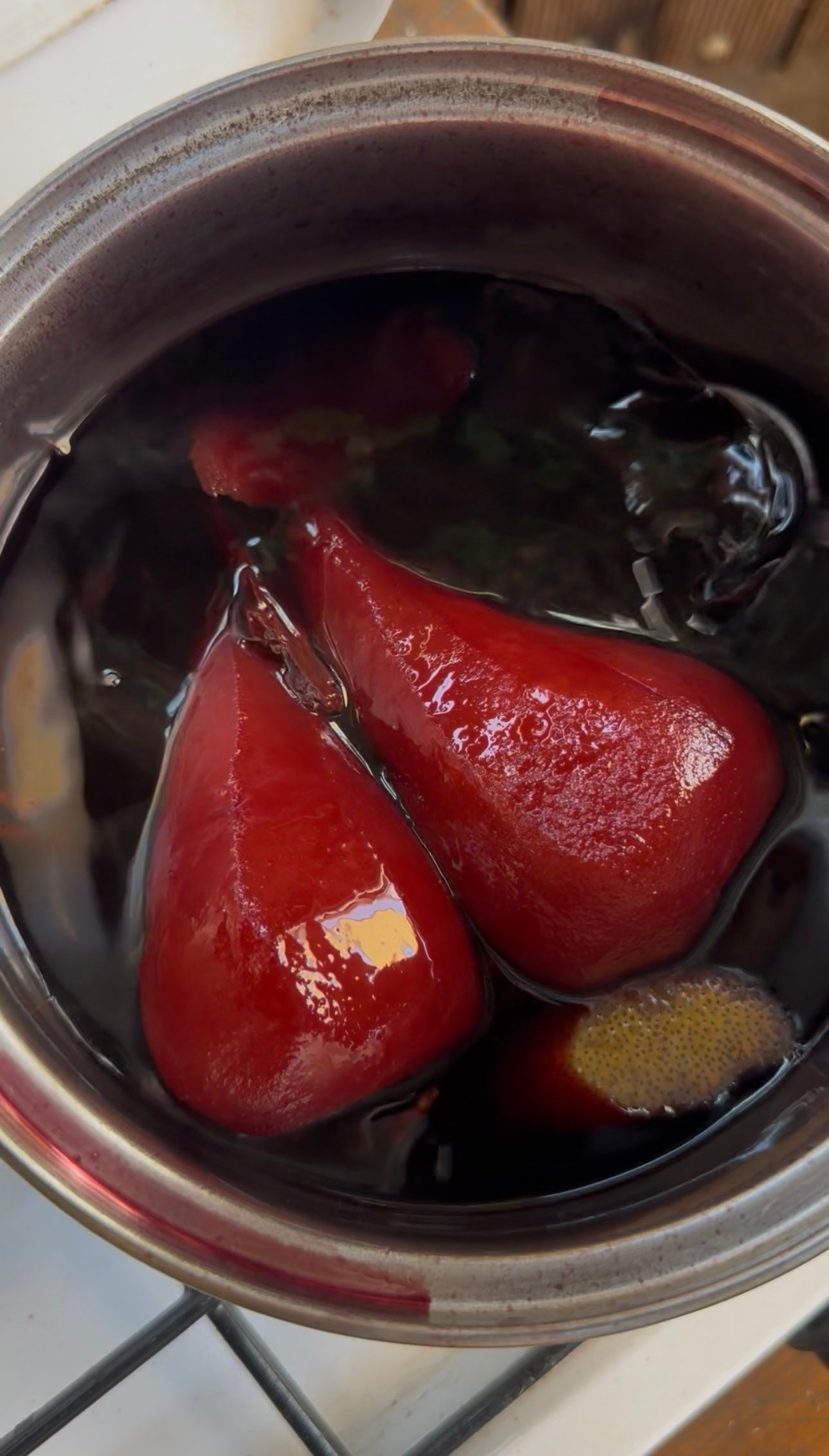 Two red poached pears sit in dark syrup inside a metal pot, with slices of orange and spices partially visible in the liquid.