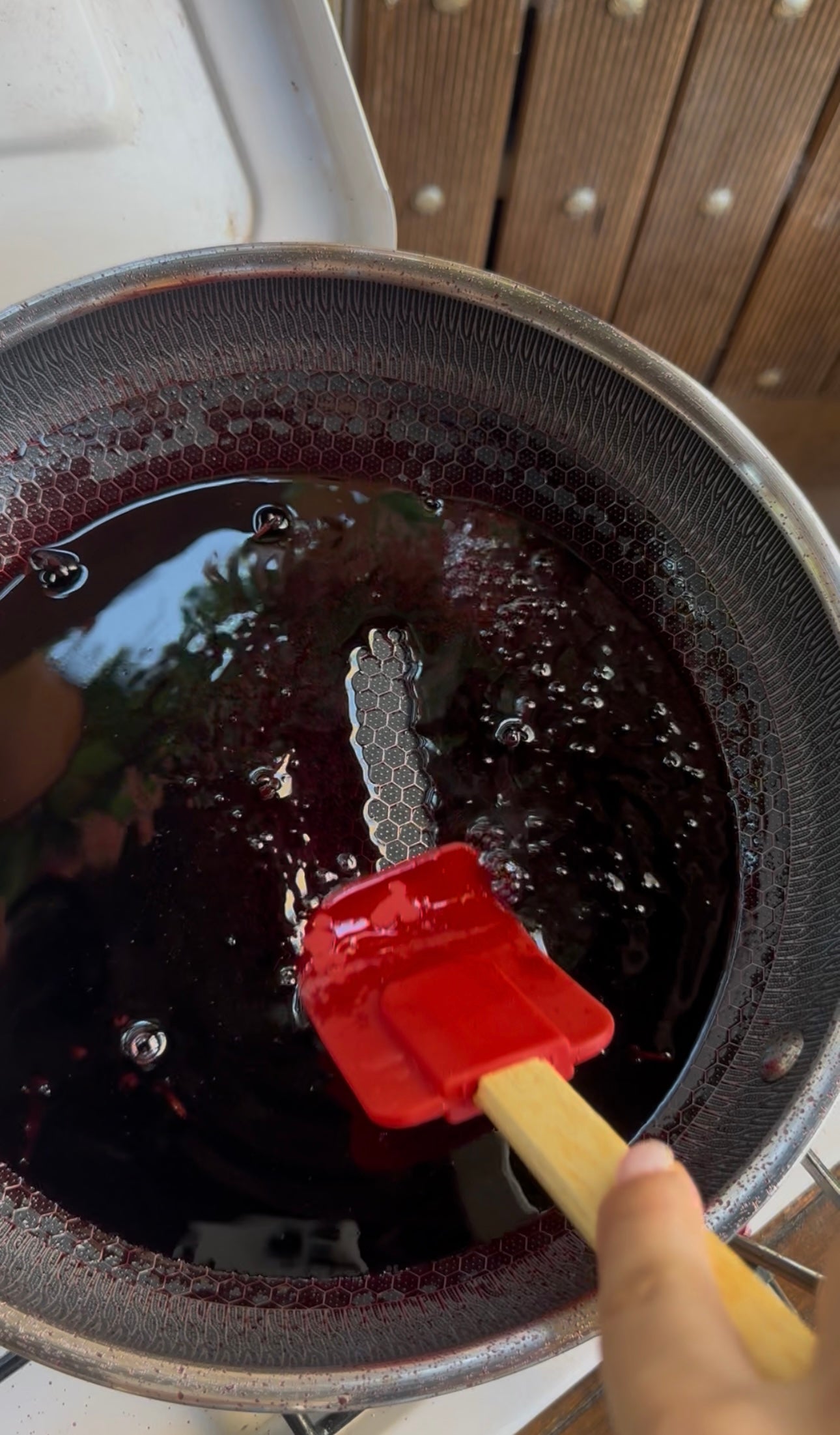 A red spatula stirs a dark liquid in a metal saucepan, creating small bubbles on the surface. The pan appears to be on a stove, with wood paneling visible in the background.