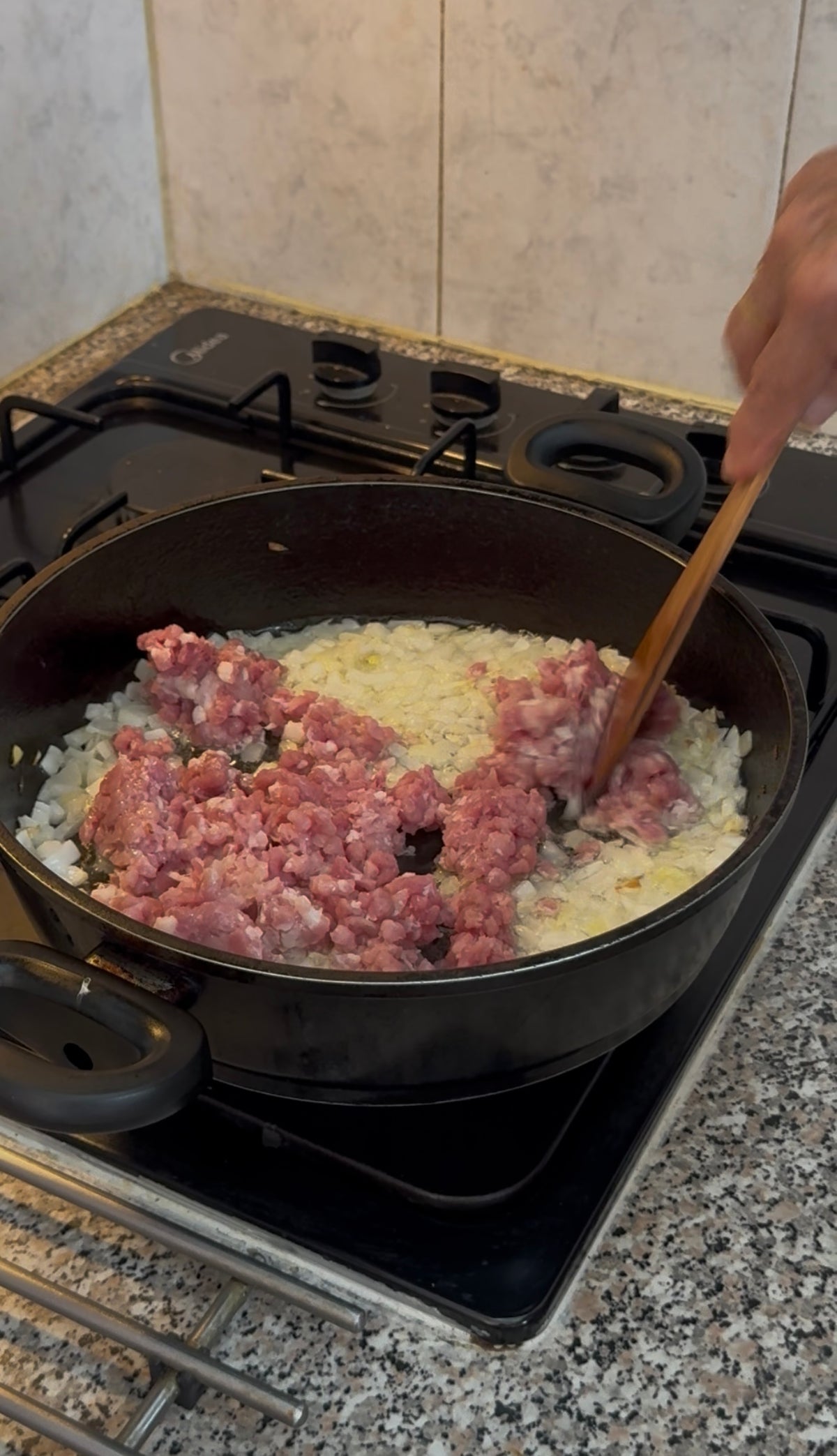 Ground meat and chopped onions are being cooked in a black pan on a stovetop. Someone is stirring the mixture with a wooden spoon. The countertop is granite, and part of the gas stove is visible.