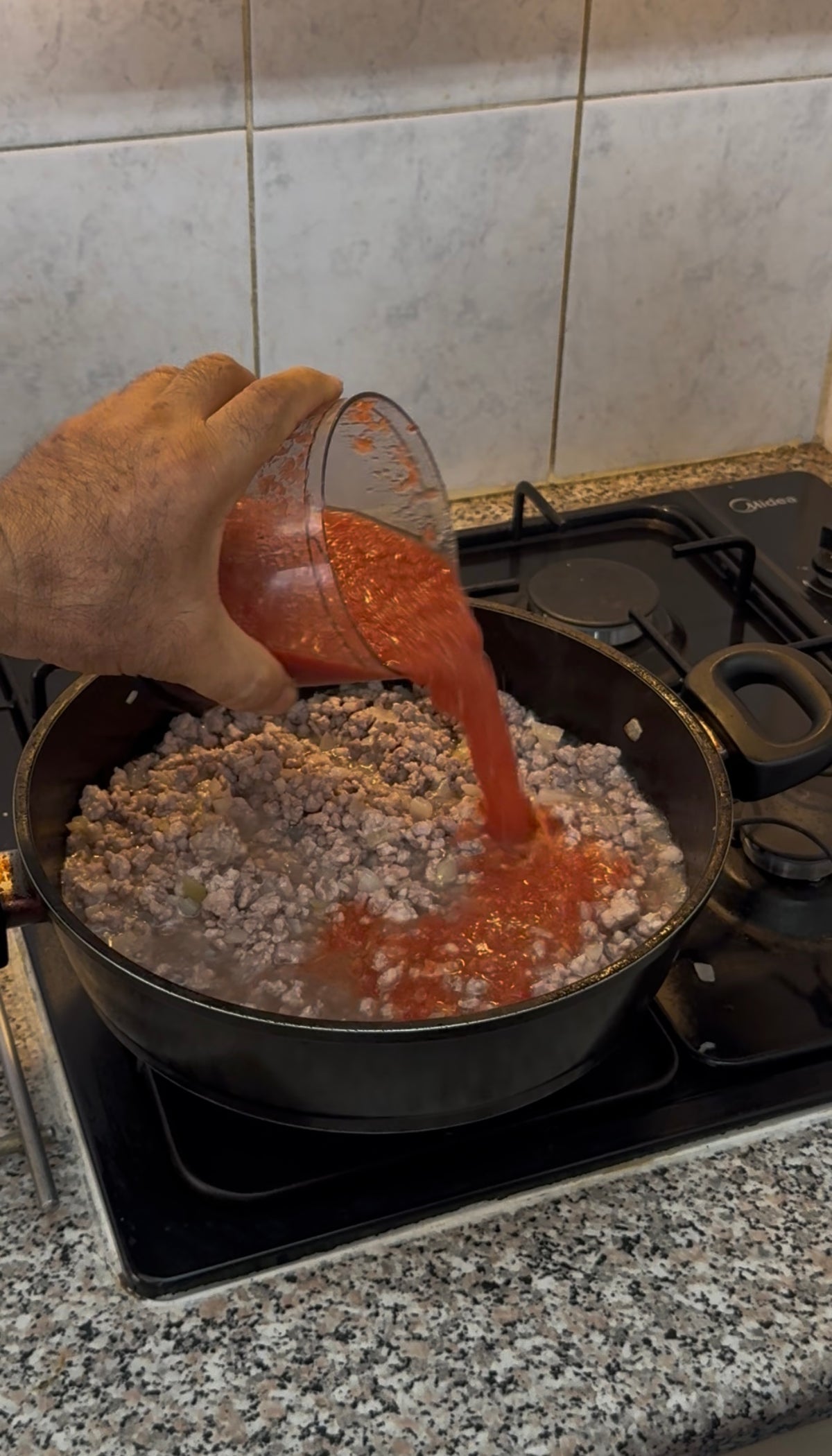 A hand pours red tomato sauce from a measuring cup into a pot of cooked ground meat on a stove.