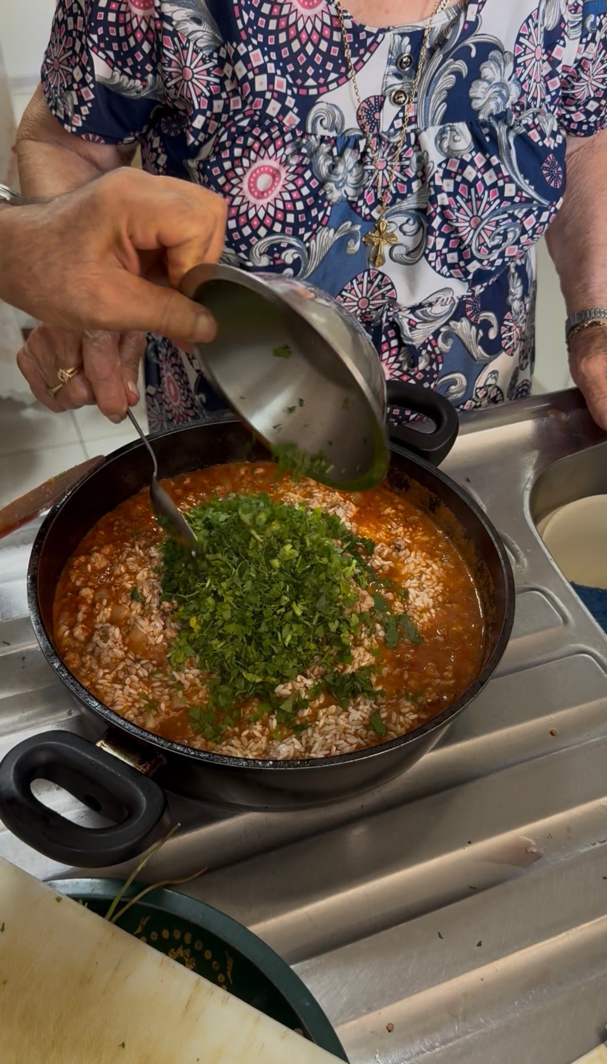 A person pours chopped herbs from a bowl into a pot of red stew on a stove. Another person stands nearby, wearing a patterned shirt.