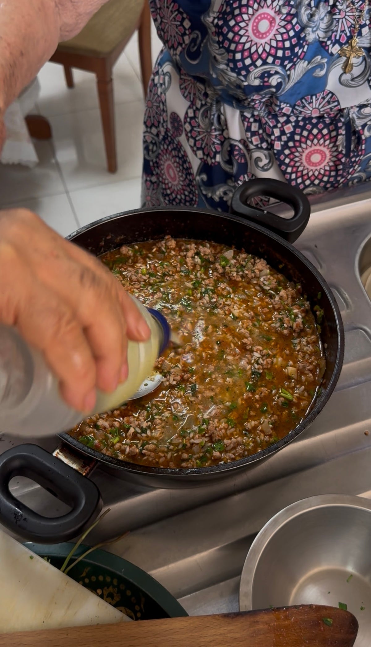 A hand pours liquid from a plastic bottle into a pot of minced meat cooking with herbs on a stove. The person wears a patterned garment.