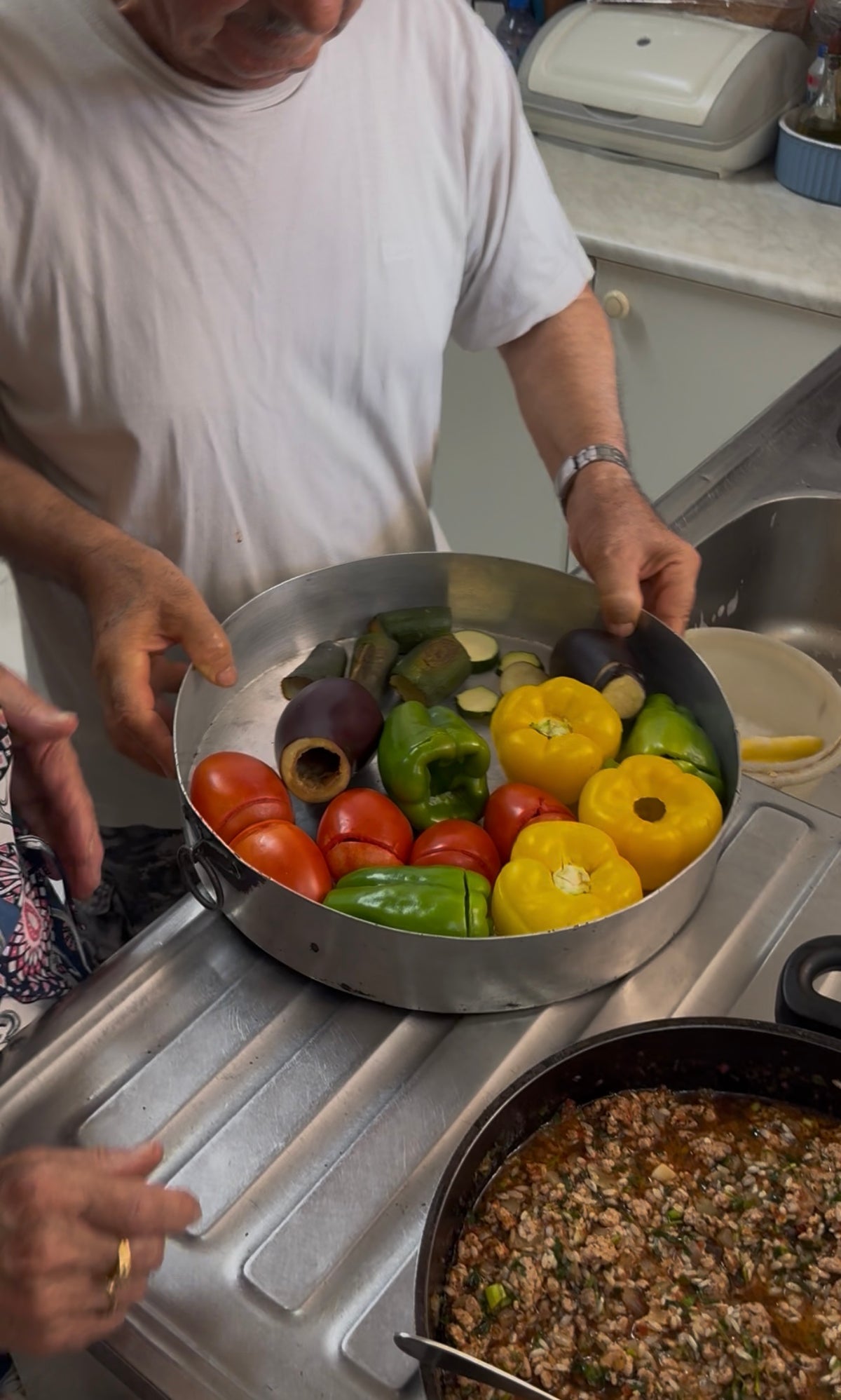 A person holds a round pan filled with hollowed tomatoes, yellow peppers, green peppers, and eggplants, preparing vegetables for stuffing in a kitchen.