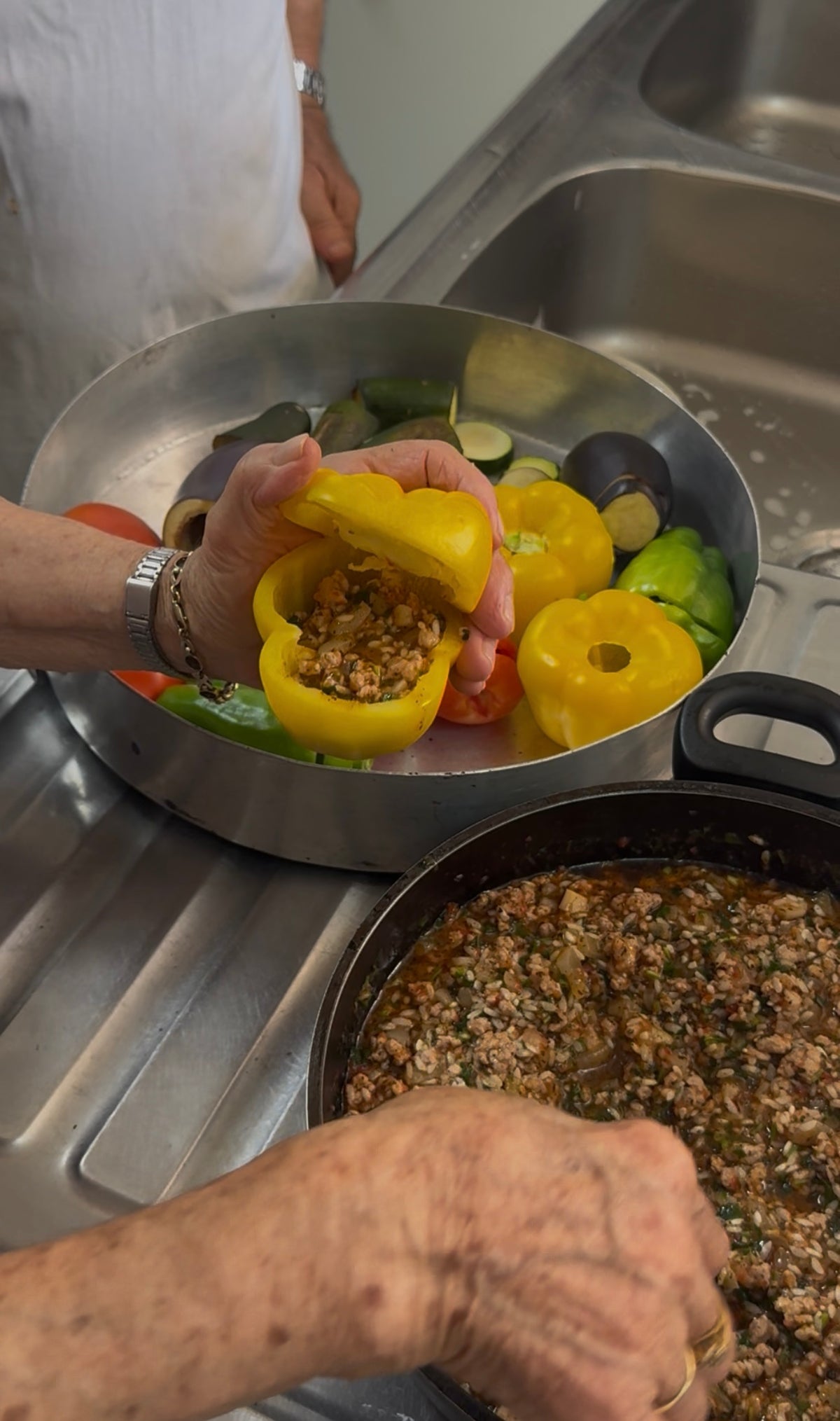 A person fills a yellow bell pepper with a ground meat mixture over a sink. Other hollowed-out vegetables and a pan of filling are nearby.