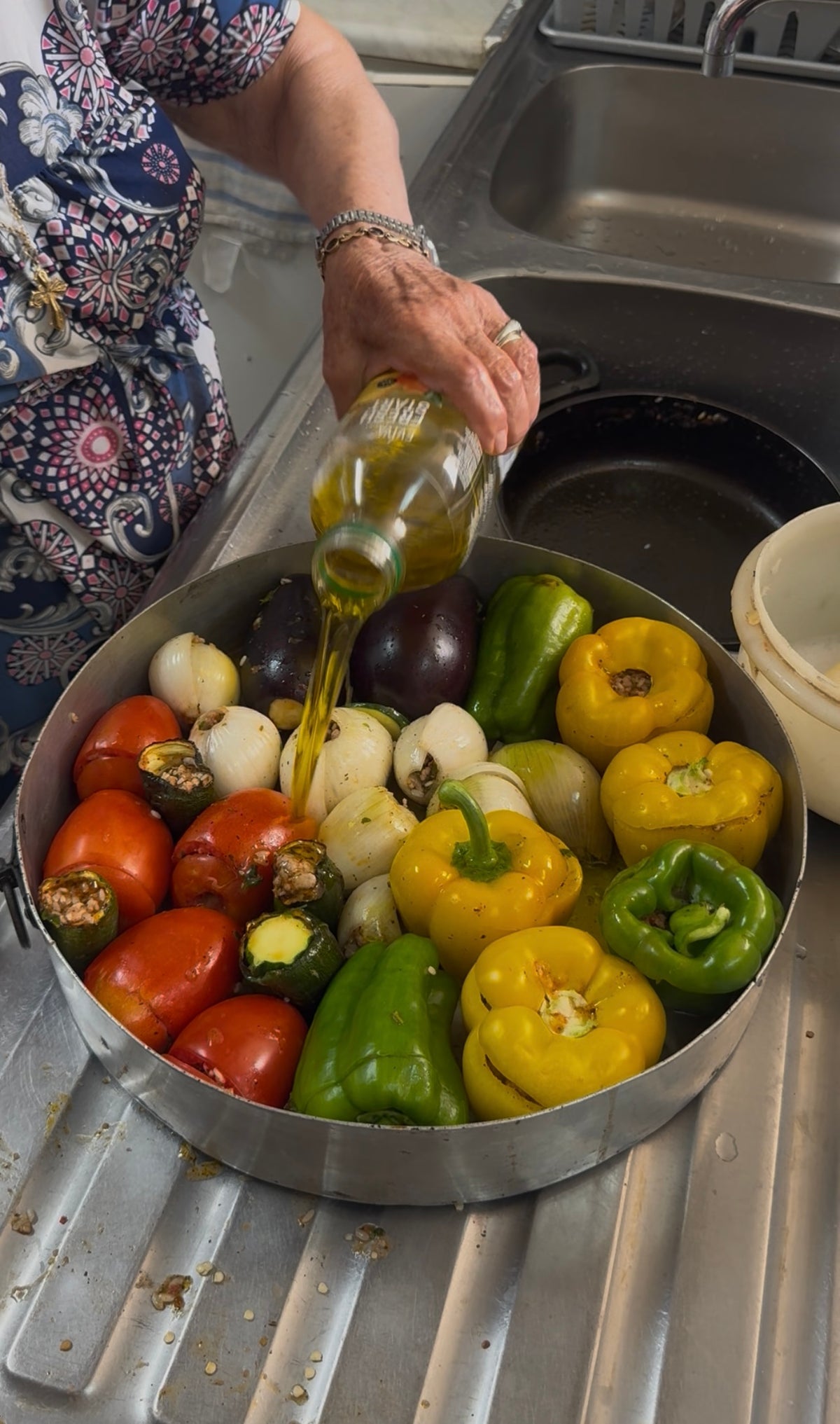 A person pours olive oil over whole tomatoes, onions, bell peppers, and eggplants arranged in a round baking pan beside a kitchen sink.