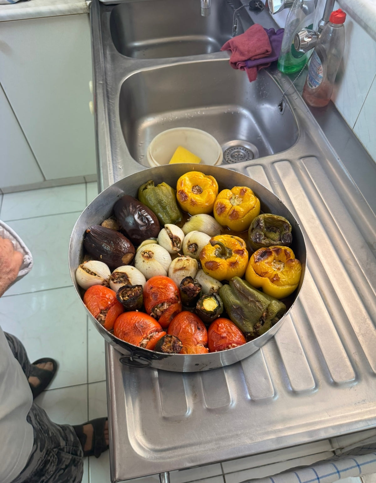 A round metal tray filled with baked vegetables, including tomatoes, bell peppers, onions, and eggplants, resting on a kitchen sink draining board. A person stands nearby.