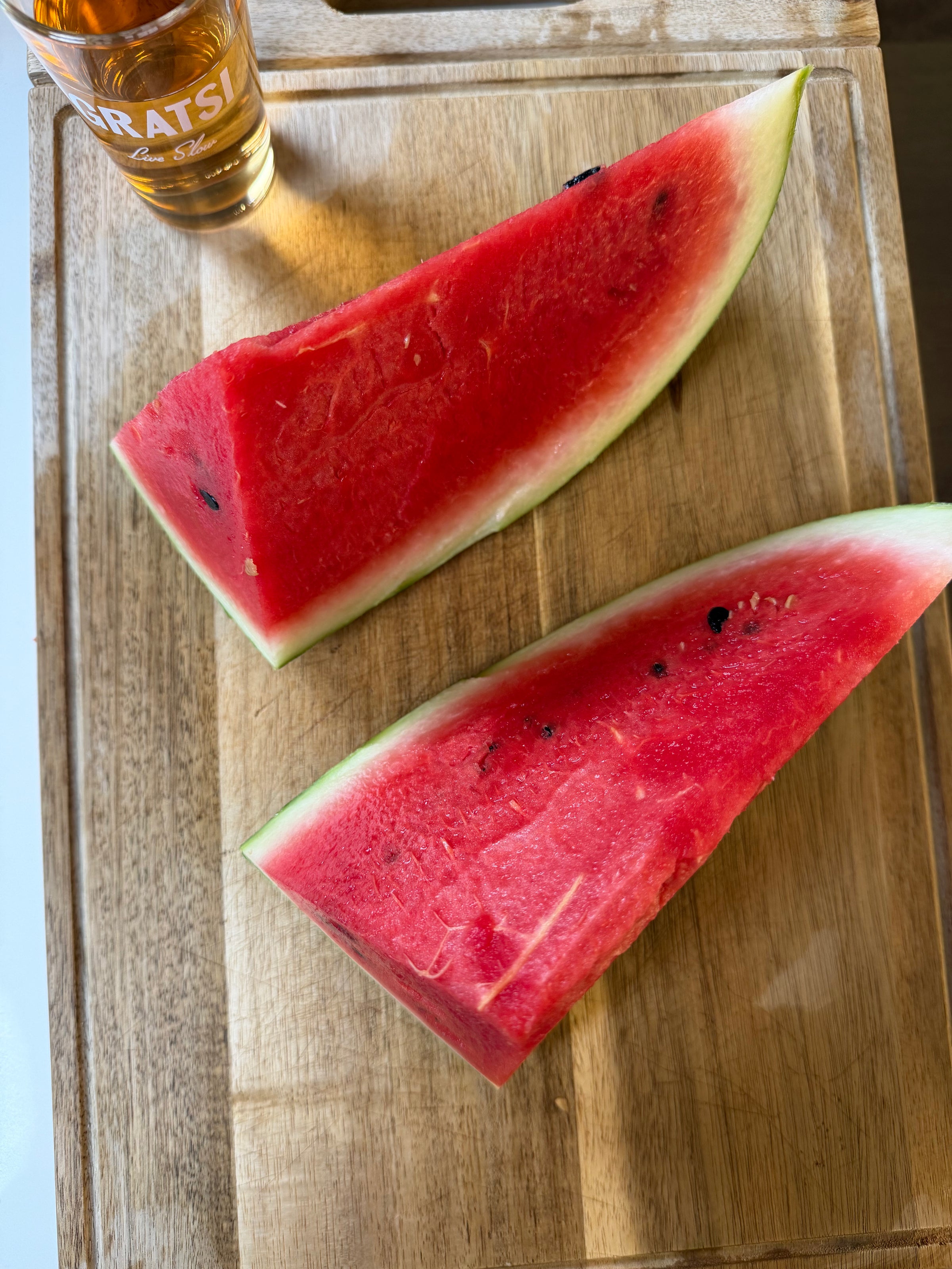 Two thick slices of watermelon on a wooden cutting board next to a small glass filled with a golden-brown liquid.