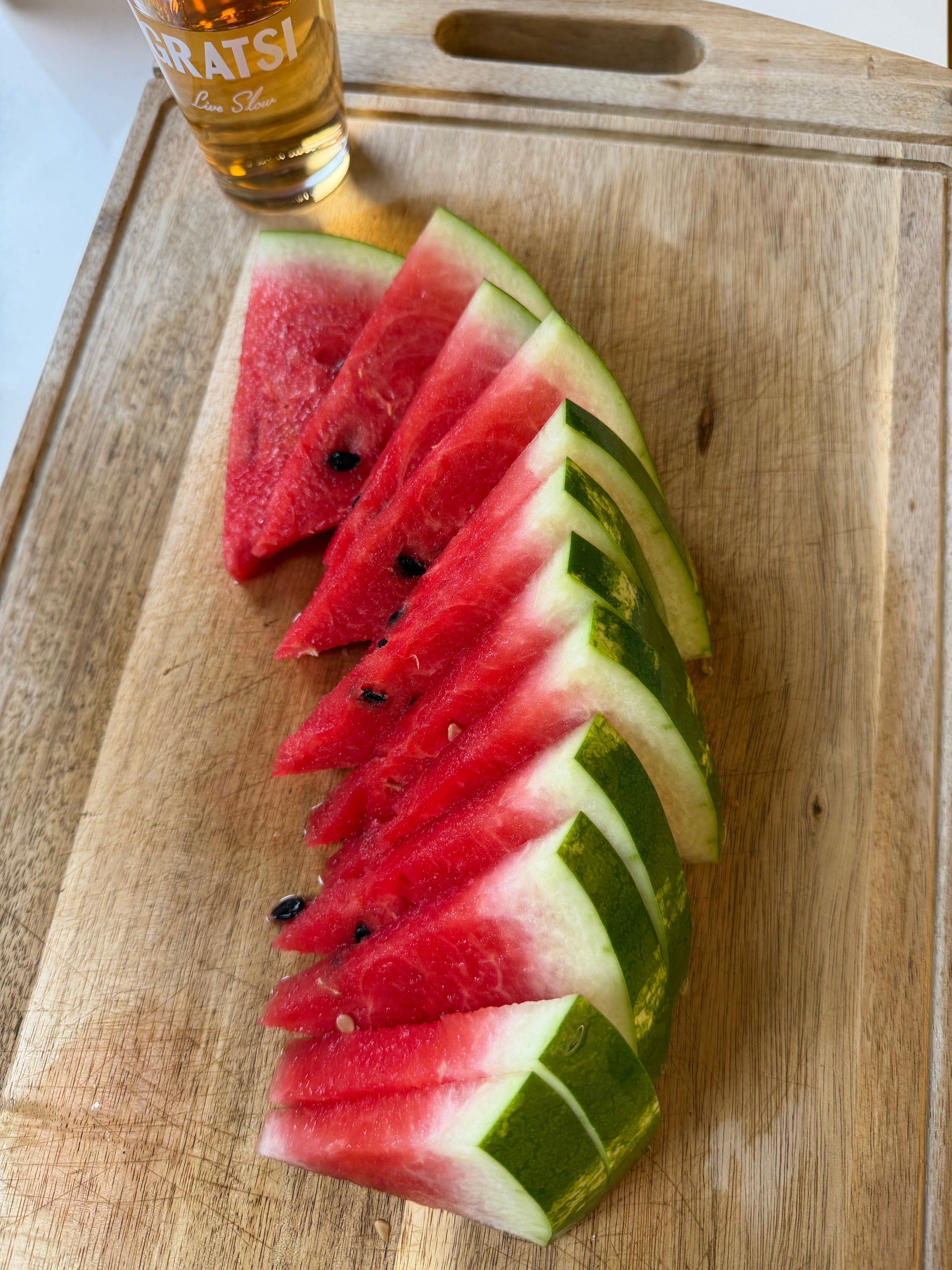 Slices of watermelon arranged in a fan shape on a wooden cutting board, with a glass of amber-colored liquid partially visible in the top left corner.