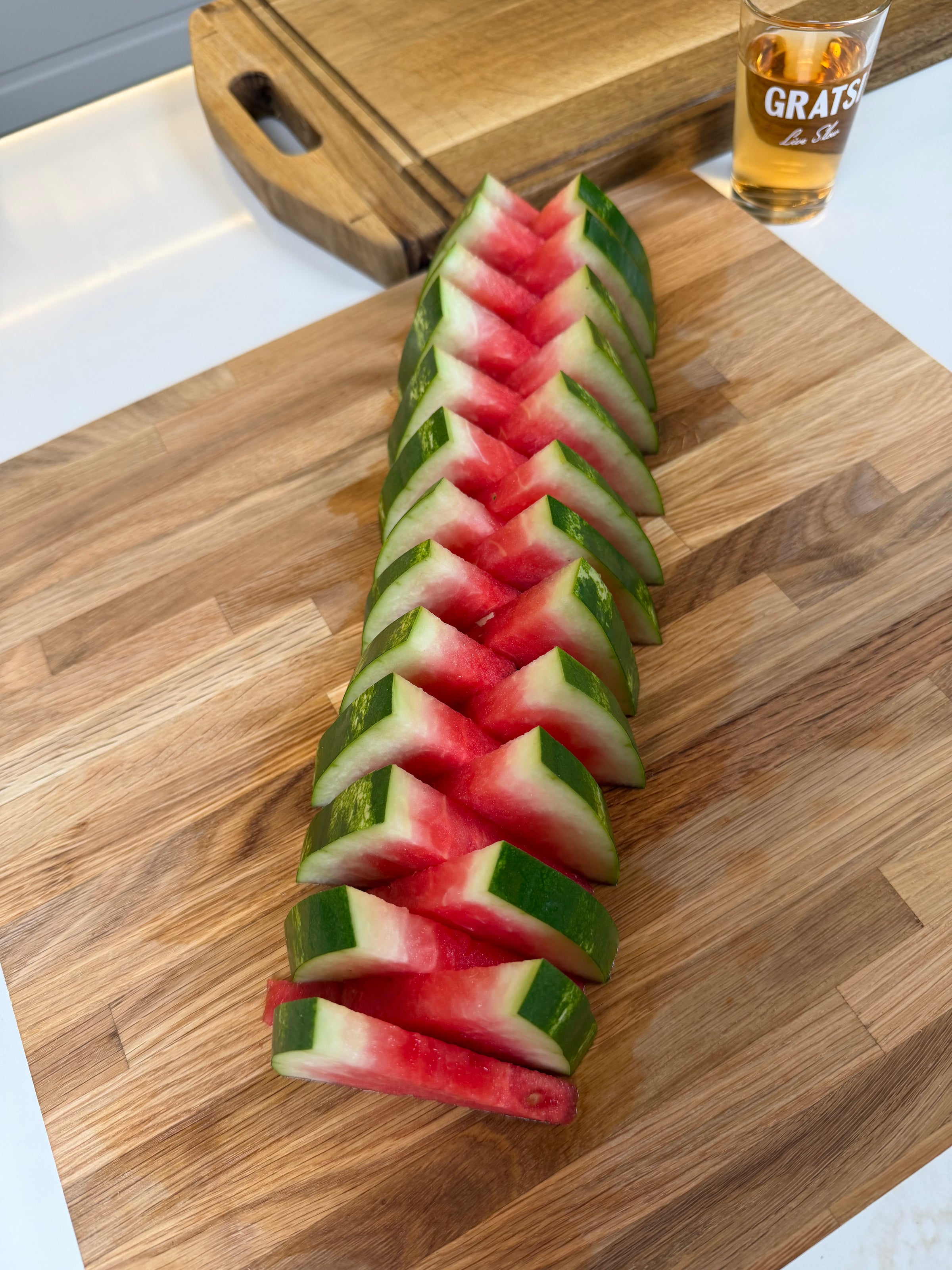 Watermelon sliced into evenly spaced triangular pieces on a wooden cutting board, with a drink in a glass labeled GRATS in the background.