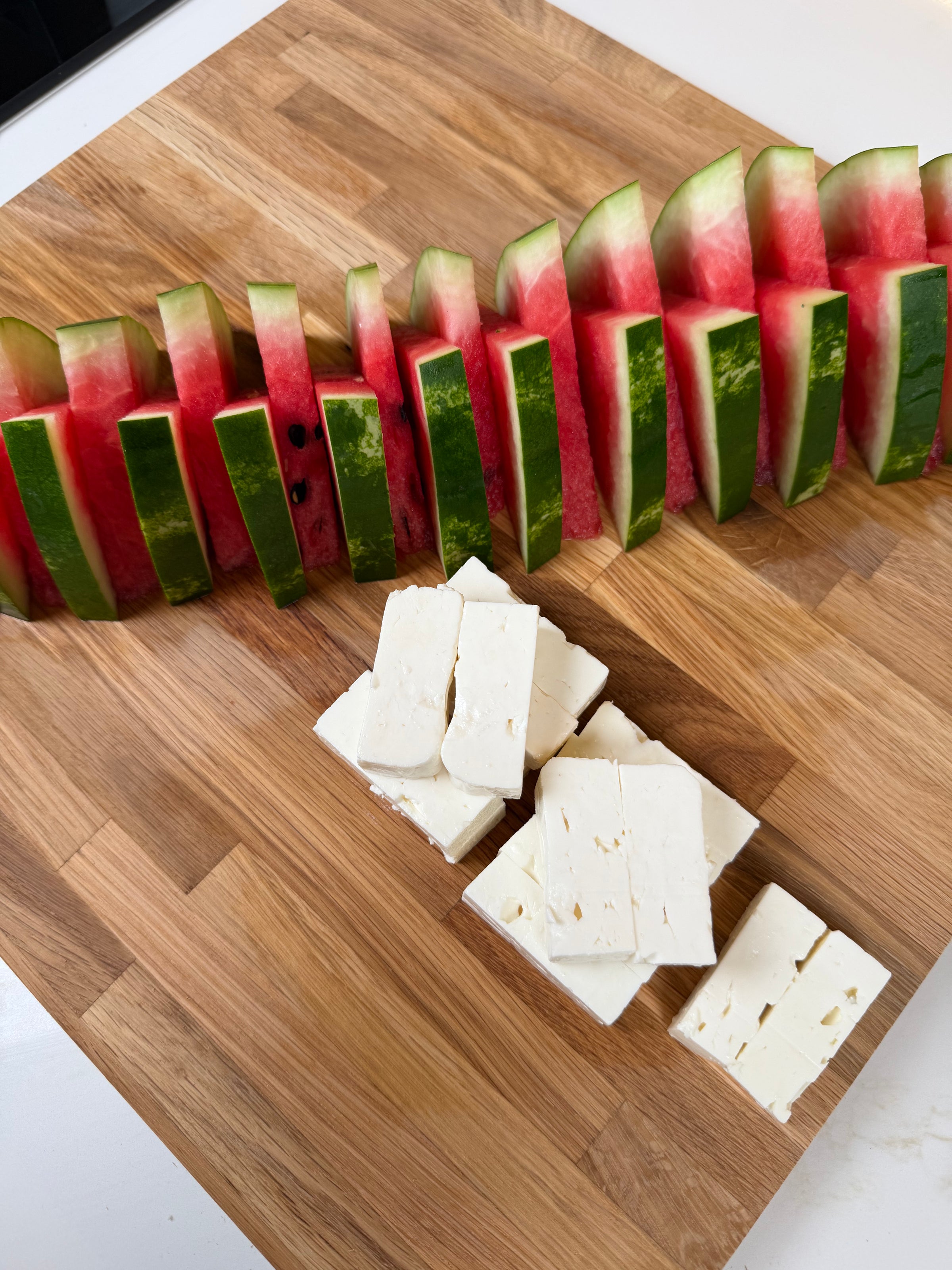 Sliced watermelon arranged in a line on a wooden cutting board, with a stack of rectangular feta cheese pieces placed nearby.