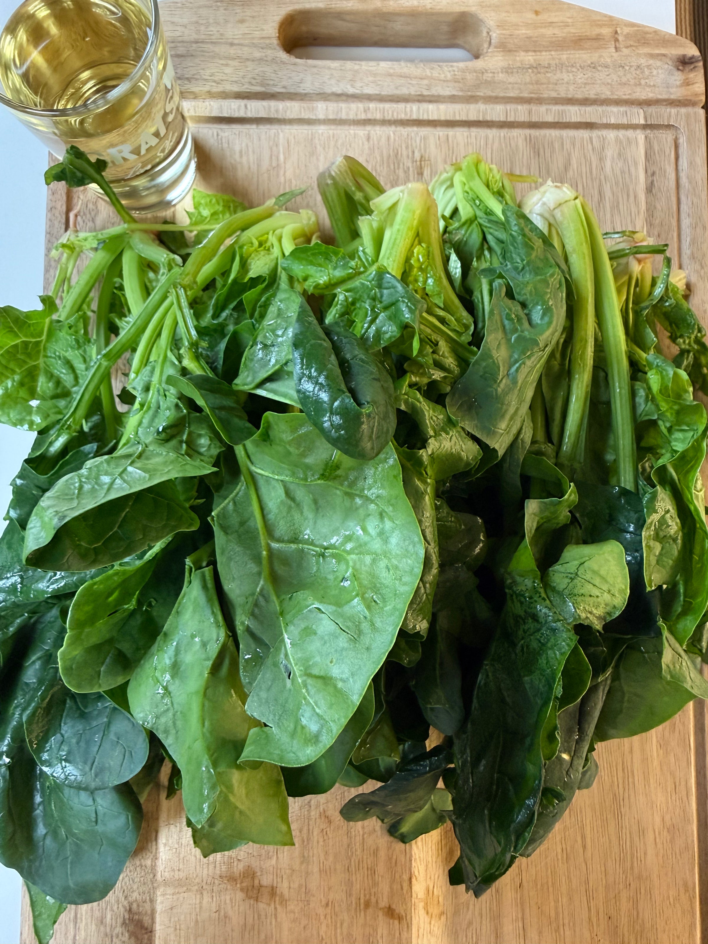 A bunch of fresh green spinach leaves on a wooden cutting board next to a glass of pale yellow liquid.