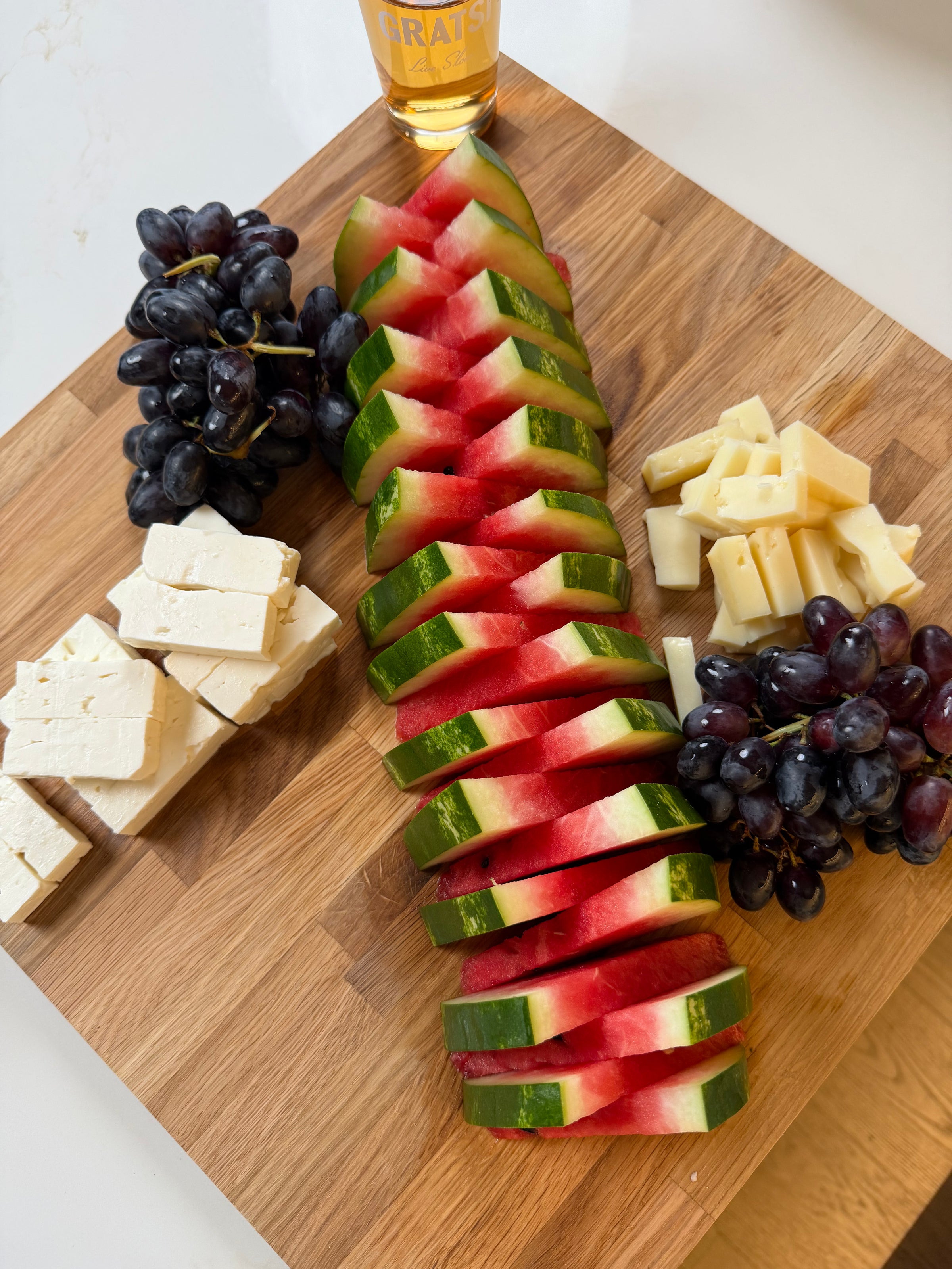 Sliced watermelon, black grapes, cubes of white and yellow cheese on a wooden board, with a drink in the background.