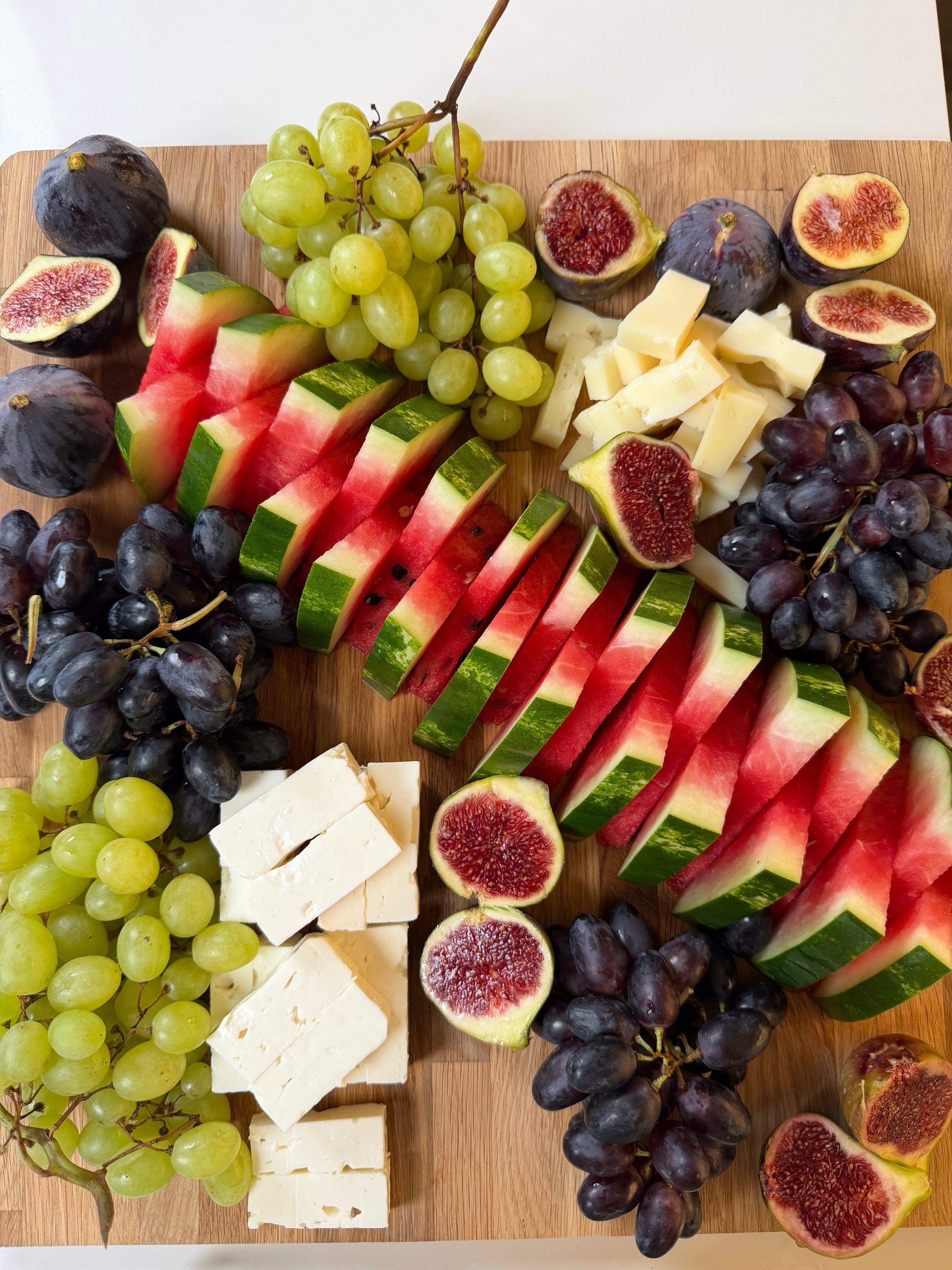 Sliced watermelon, figs, green and purple grapes, and cubes of white and yellow cheese arranged on a wooden board.