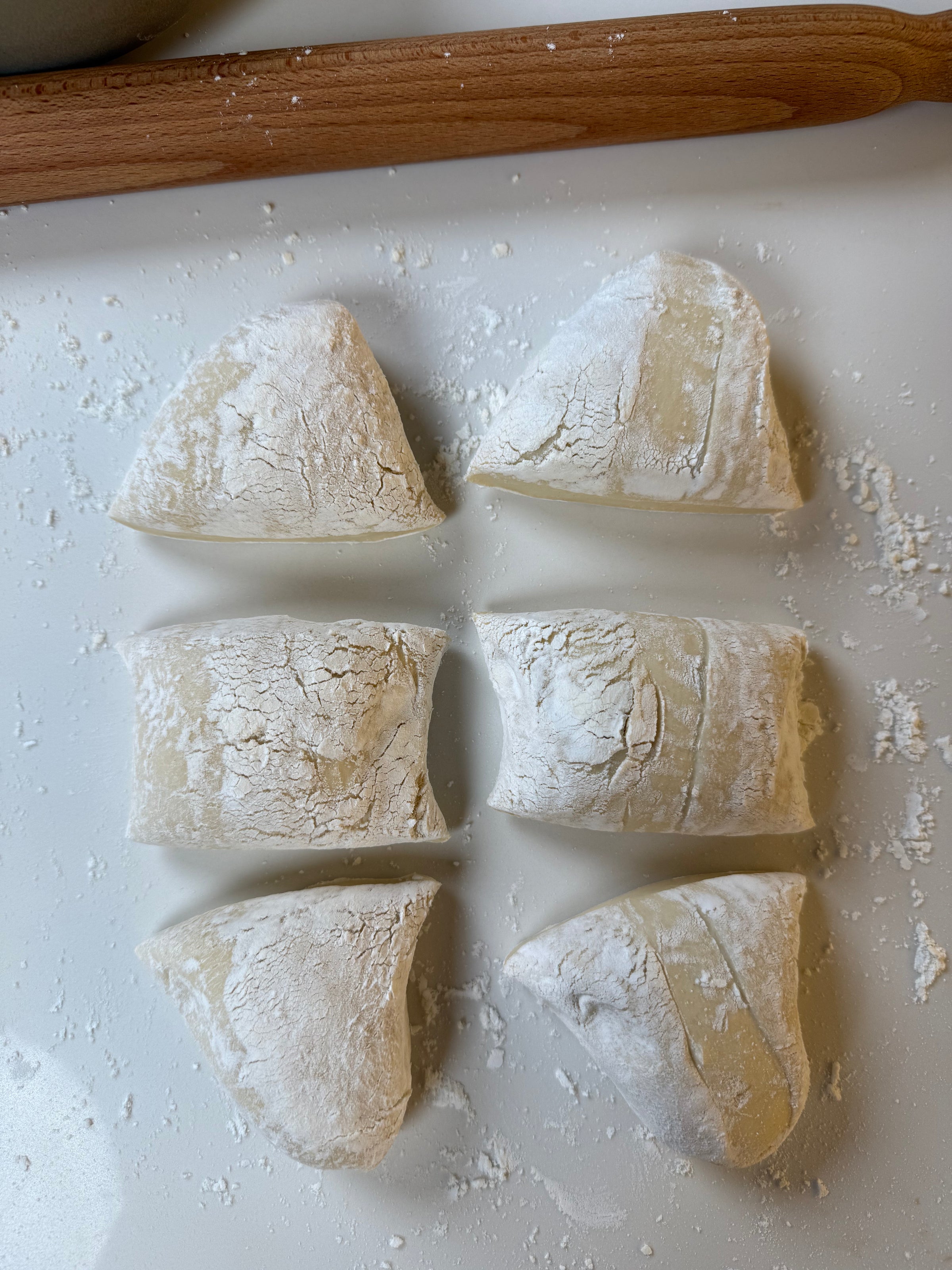 Six pieces of dough, dusted with flour, are arranged on a white surface. A wooden rolling pin is partially visible at the top of the image.