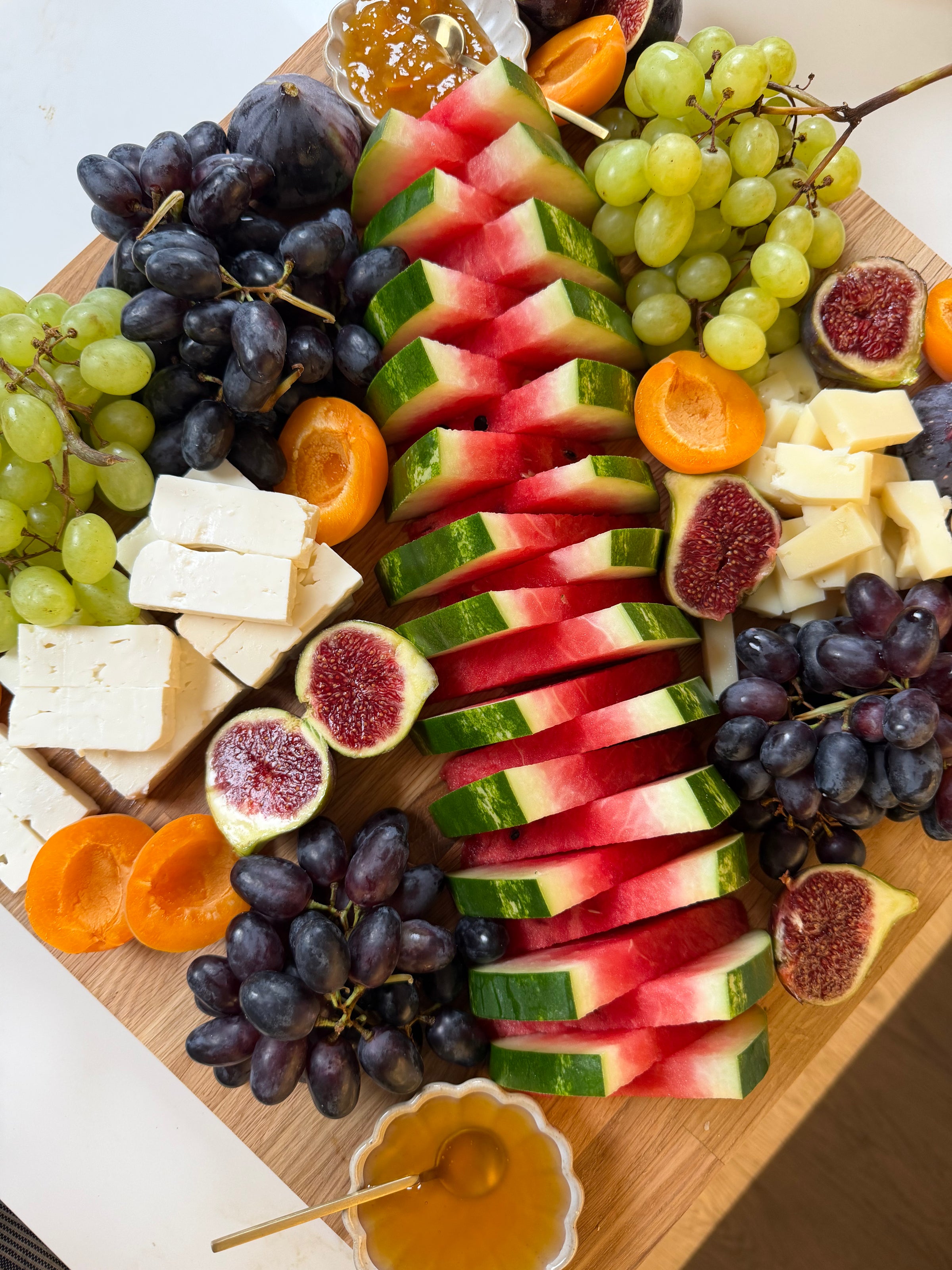 A wooden board with sliced watermelon, green and purple grapes, figs, apricots, chunks of cheese, and small bowls of honey and jam.