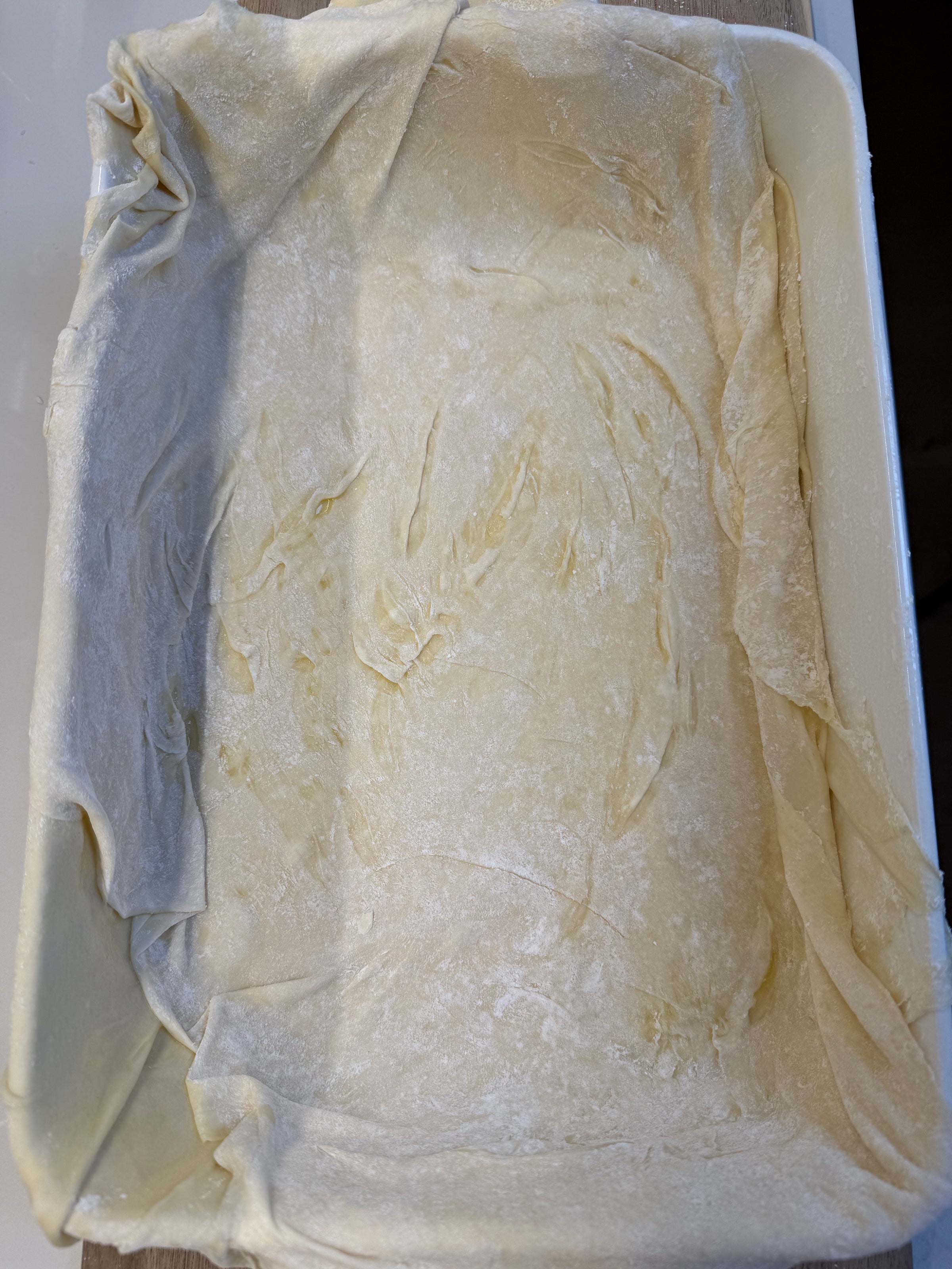 A rectangular baking dish lined with thin, slightly wrinkled sheets of phyllo dough, lightly dusted with flour and brushed with oil.