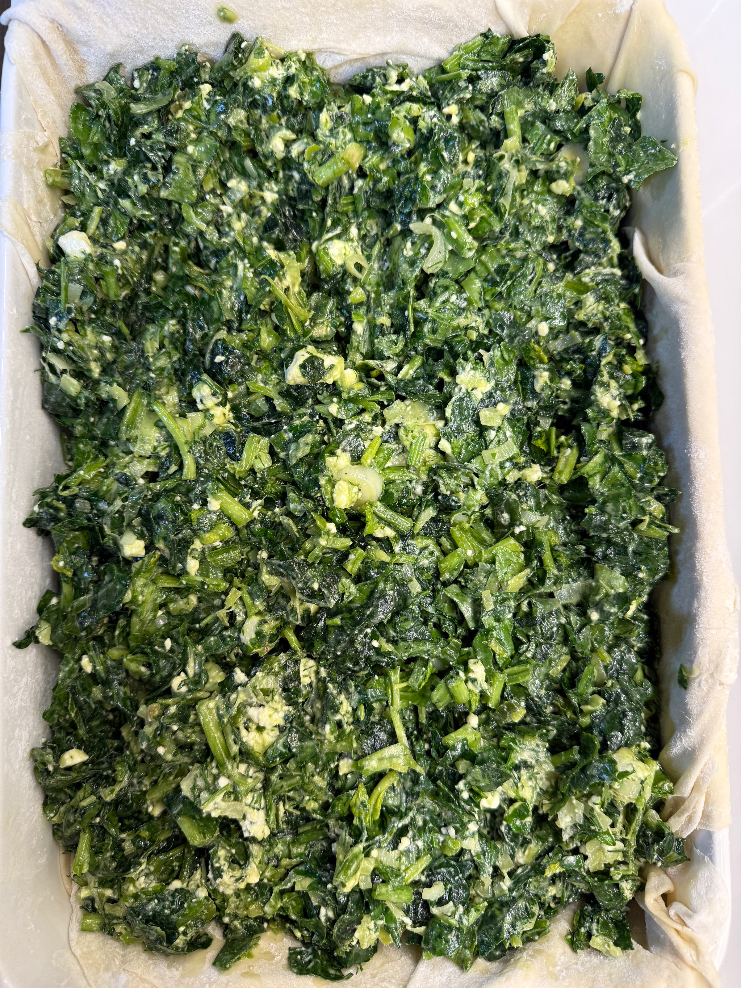 A close-up of a baking dish lined with pastry and filled with a mixture of chopped spinach, herbs, and cheese, ready to be baked.